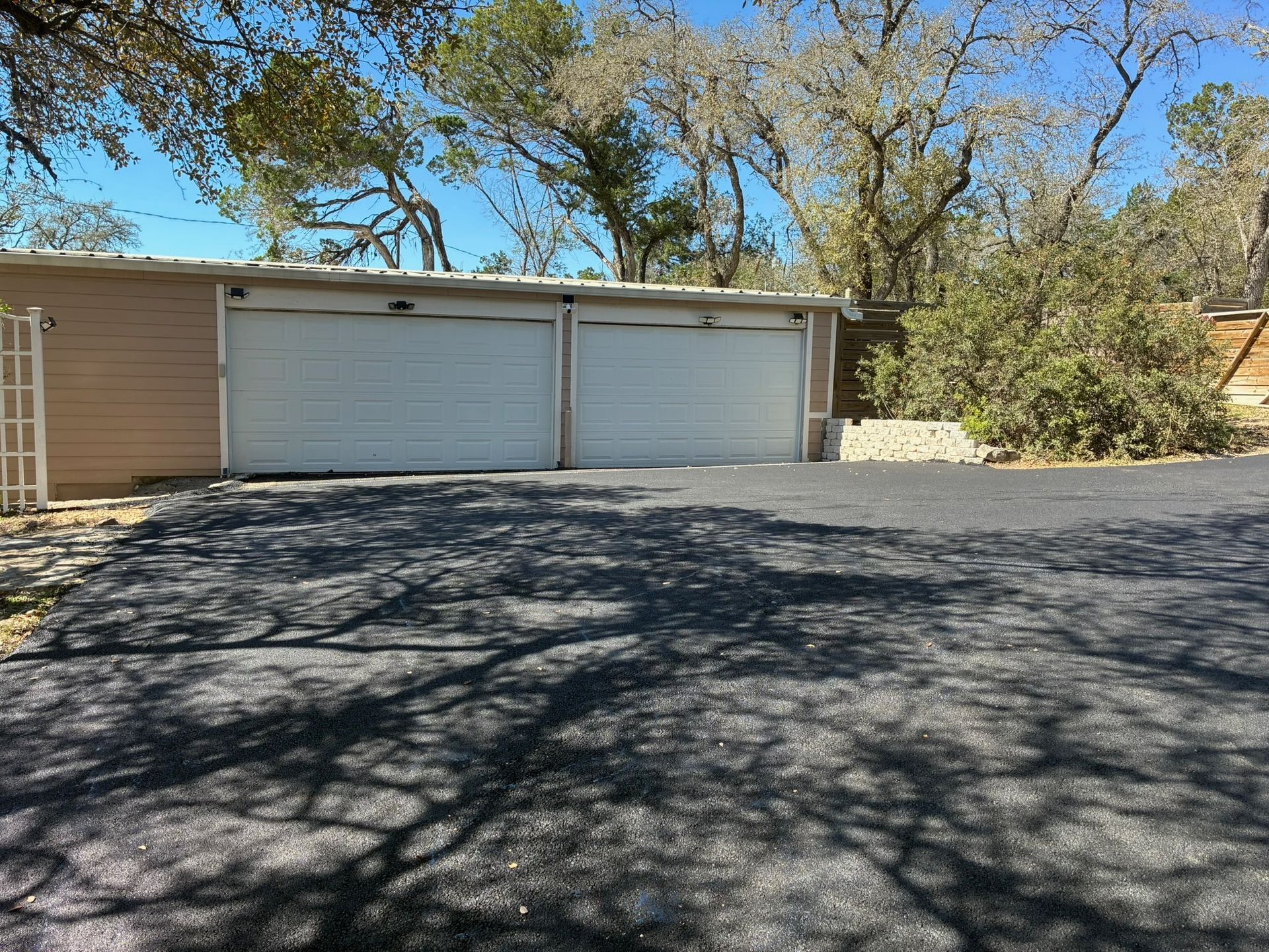 A tan two-car garage with white doors sits at the end of a paved driveway, shaded by dappled light from overhead trees.