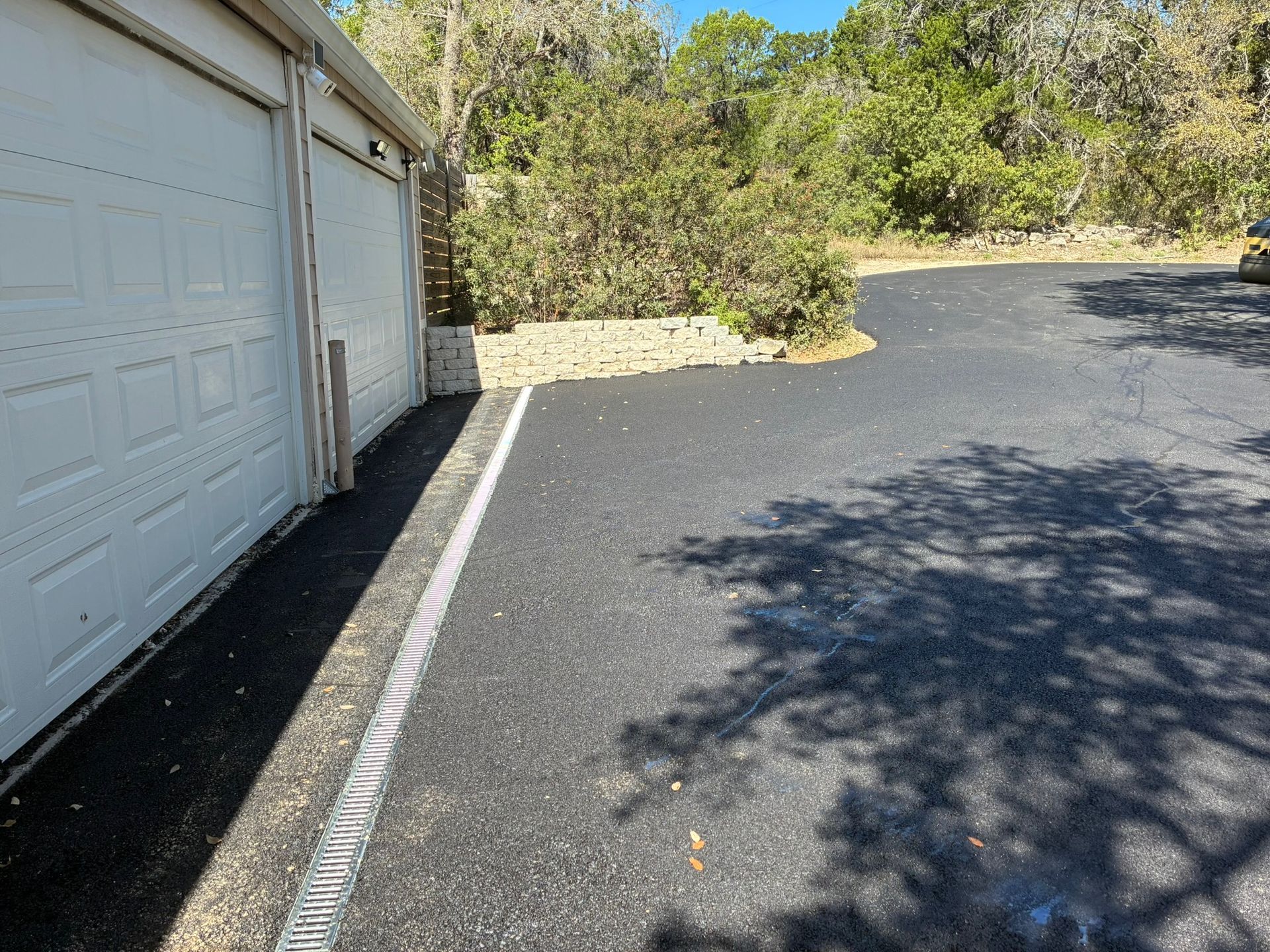 A paved driveway leads to a closed two-car garage next to a landscaped garden bed with trees in the background.