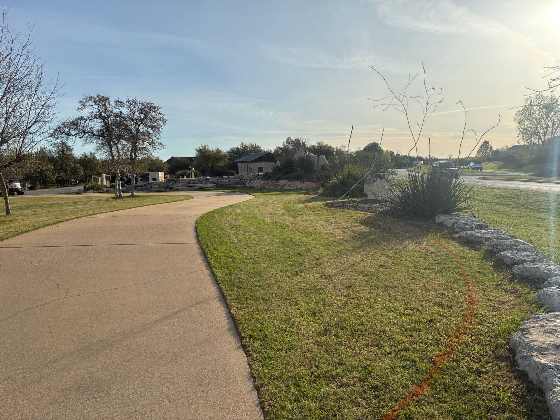 A paved walking path winds through a grassy park area with landscaping rocks and trees under a clear sky.