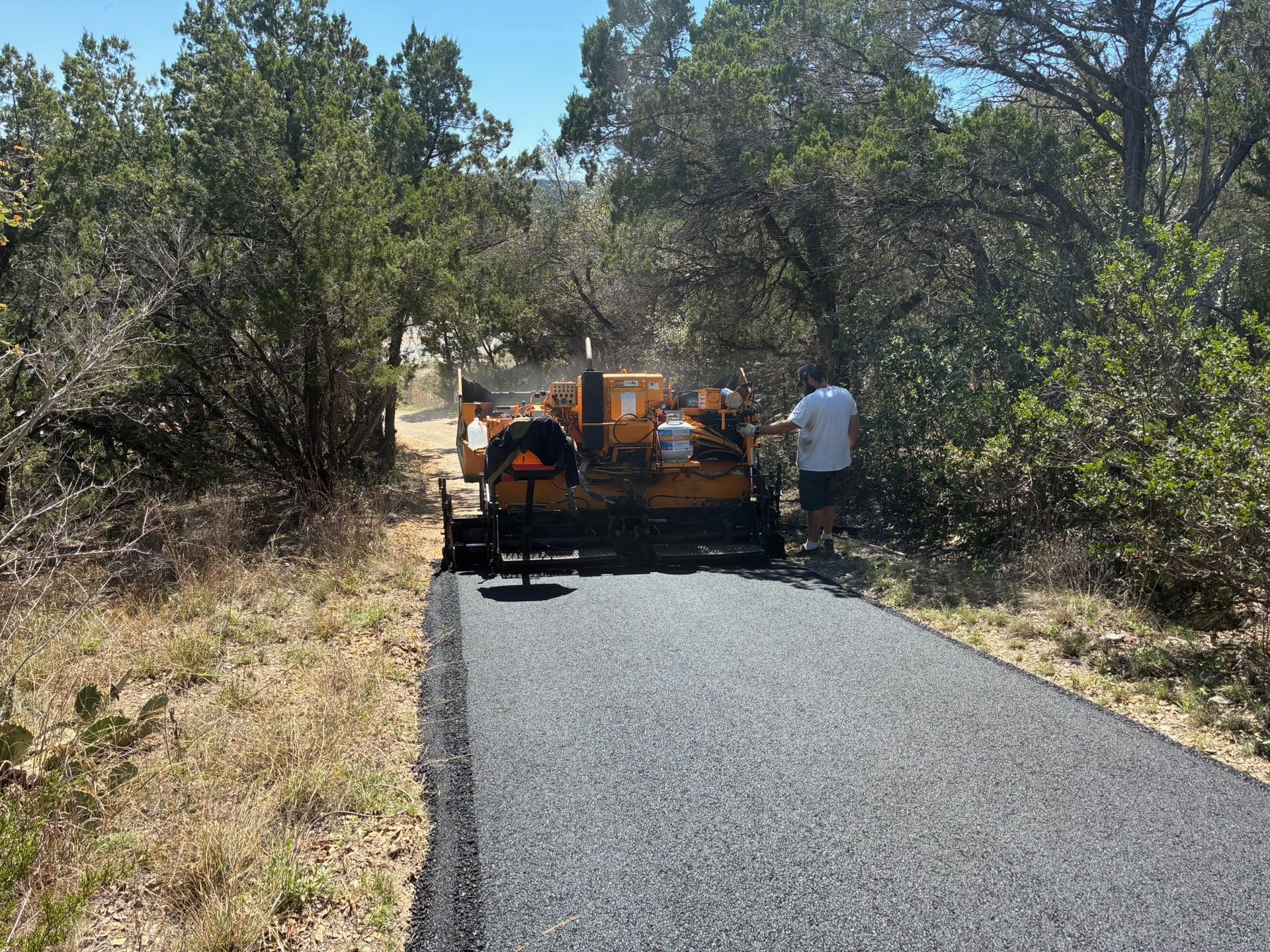 A yellow asphalt paver moves along a narrow road being newly paved through a wooded, sunny landscape.