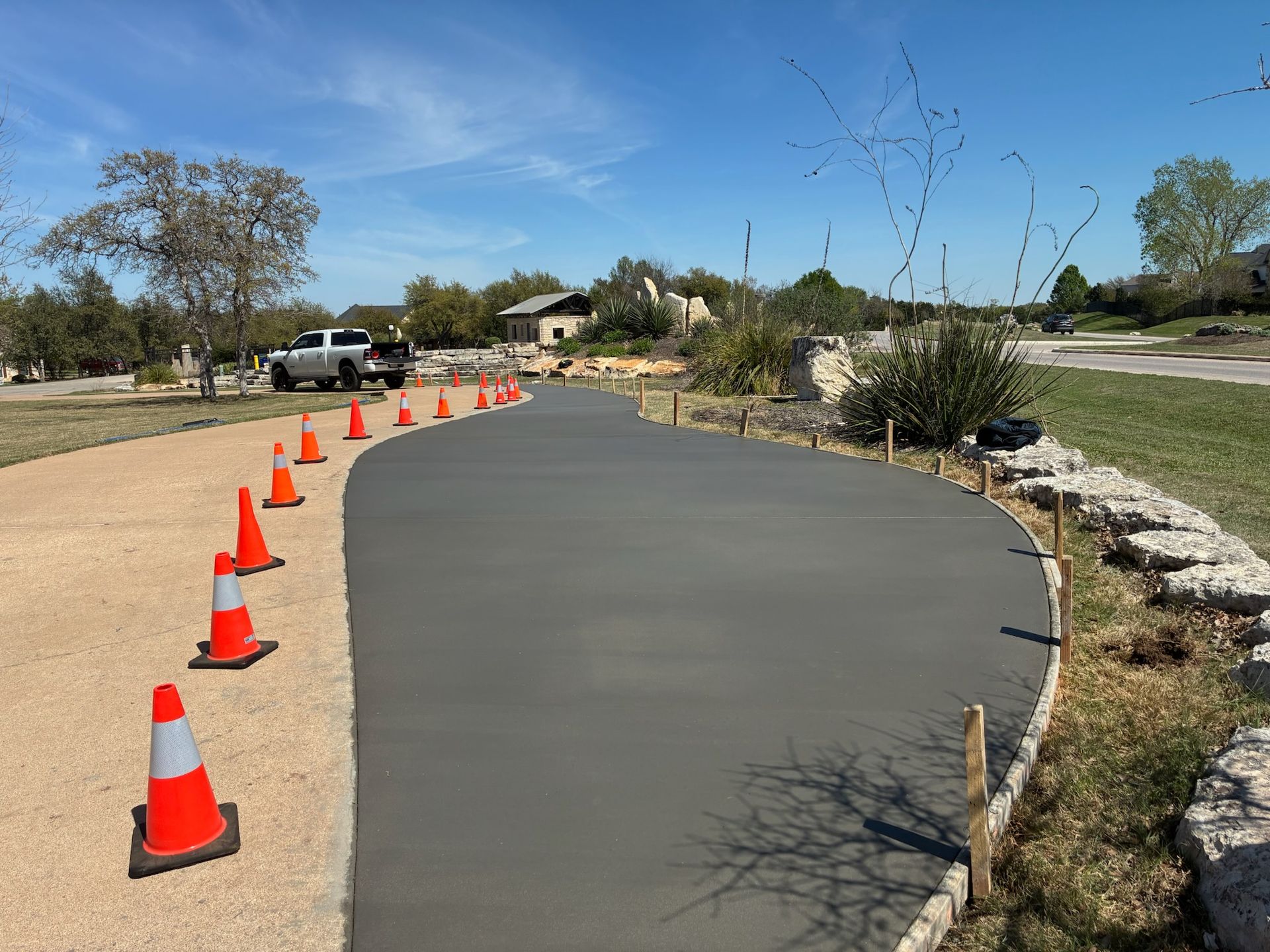 A newly poured gray concrete walkway curves through a landscape, lined with orange traffic cones and stone edging.