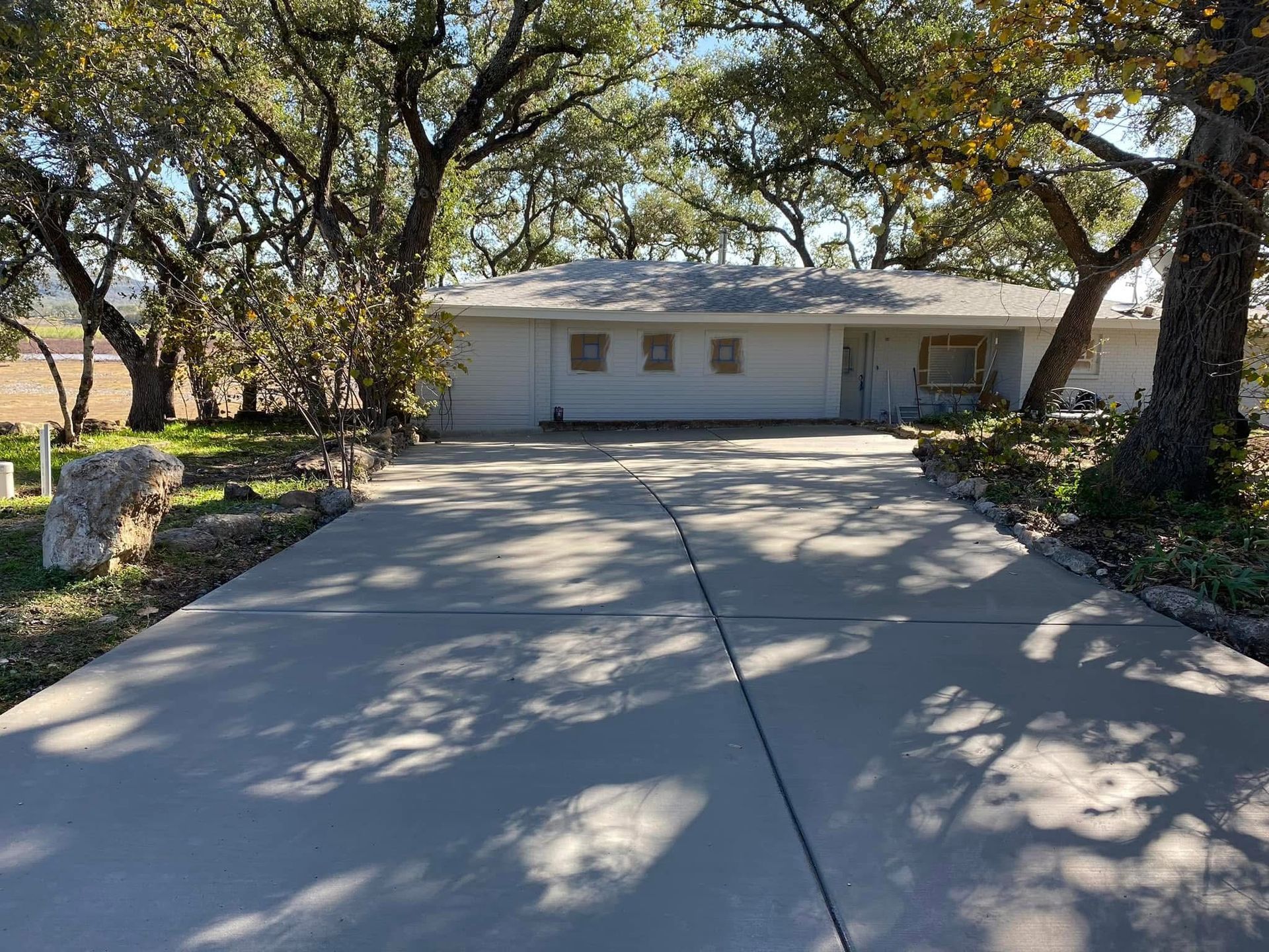 A single-story, light-colored ranch house sits at the end of a paved driveway, surrounded by many large, leafy trees.