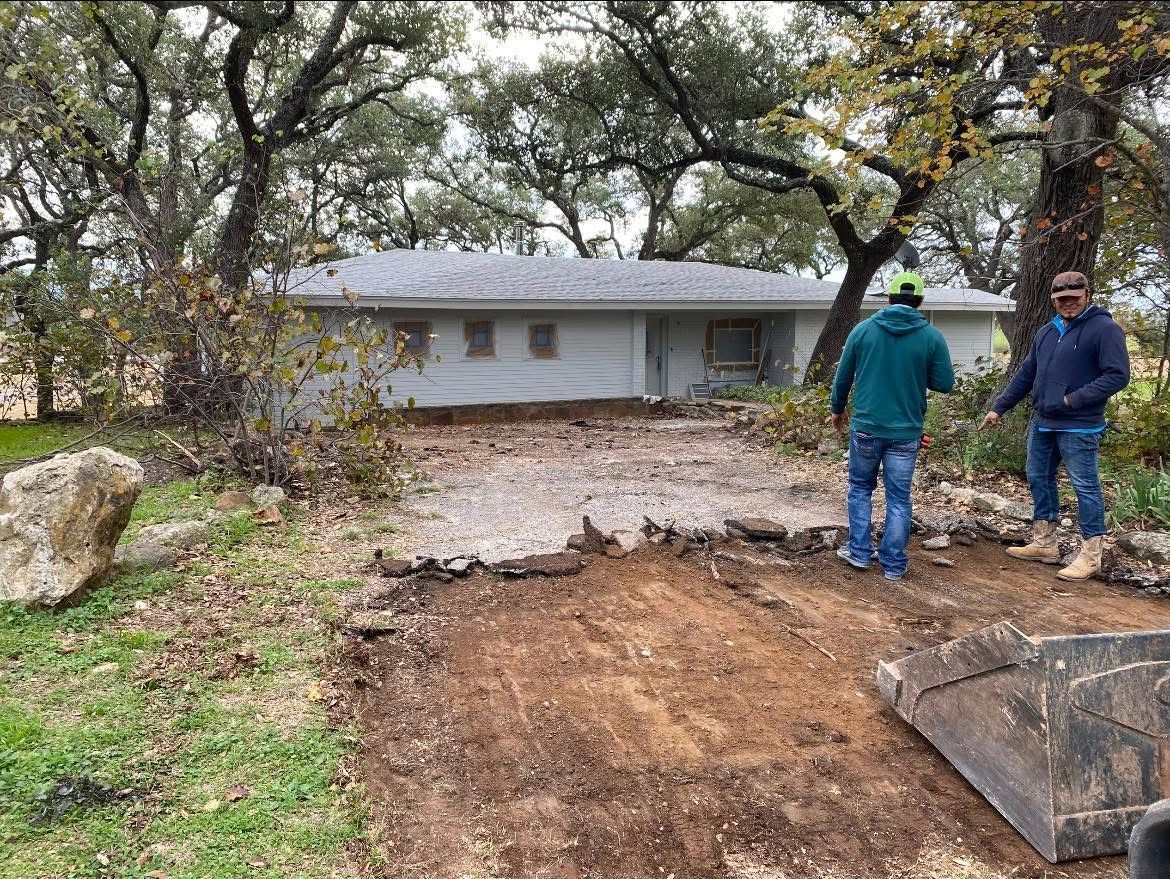 Two people stand outdoors near a freshly excavated dirt area in front of a house, with trees and a large stone nearby.
