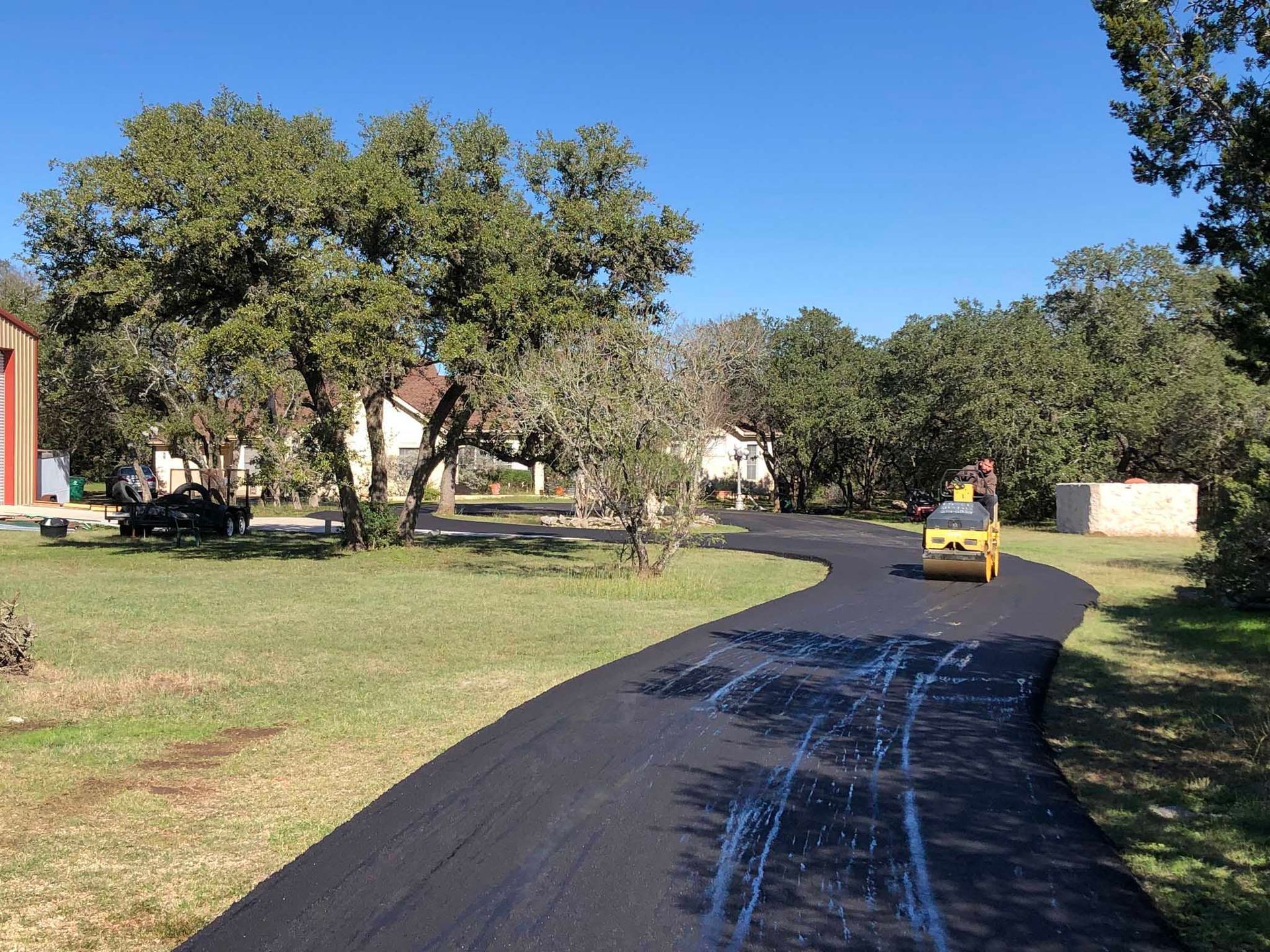 Asphalt paving a curved driveway with a roller machine; green grass, trees, and houses in the background.