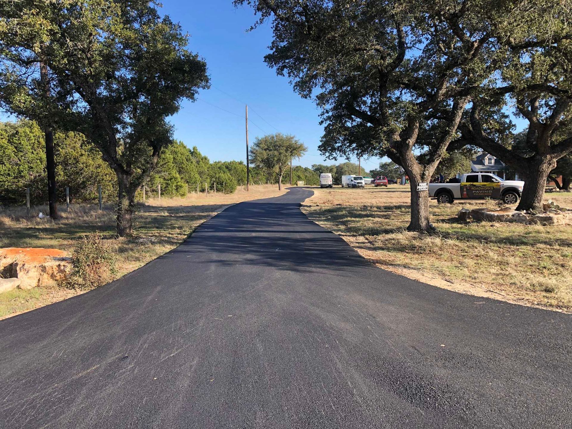 Asphalt road through a rural landscape, flanked by trees under a blue sky.