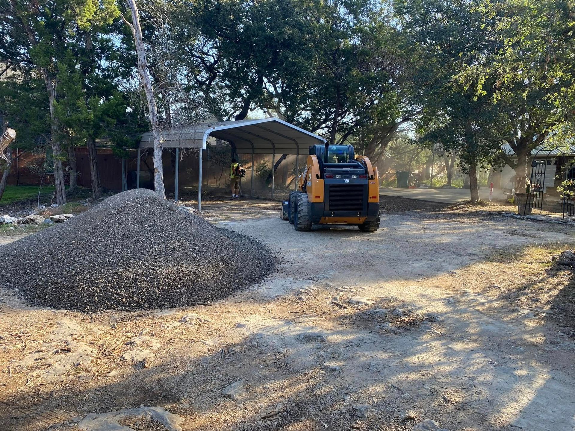 A skid steer moves gravel near a shed, trees, and a large pile of gravel outdoors.