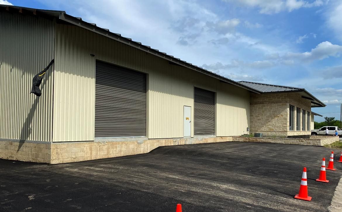 Tan building with roll-up doors, black asphalt, and orange cones. Blue sky visible.