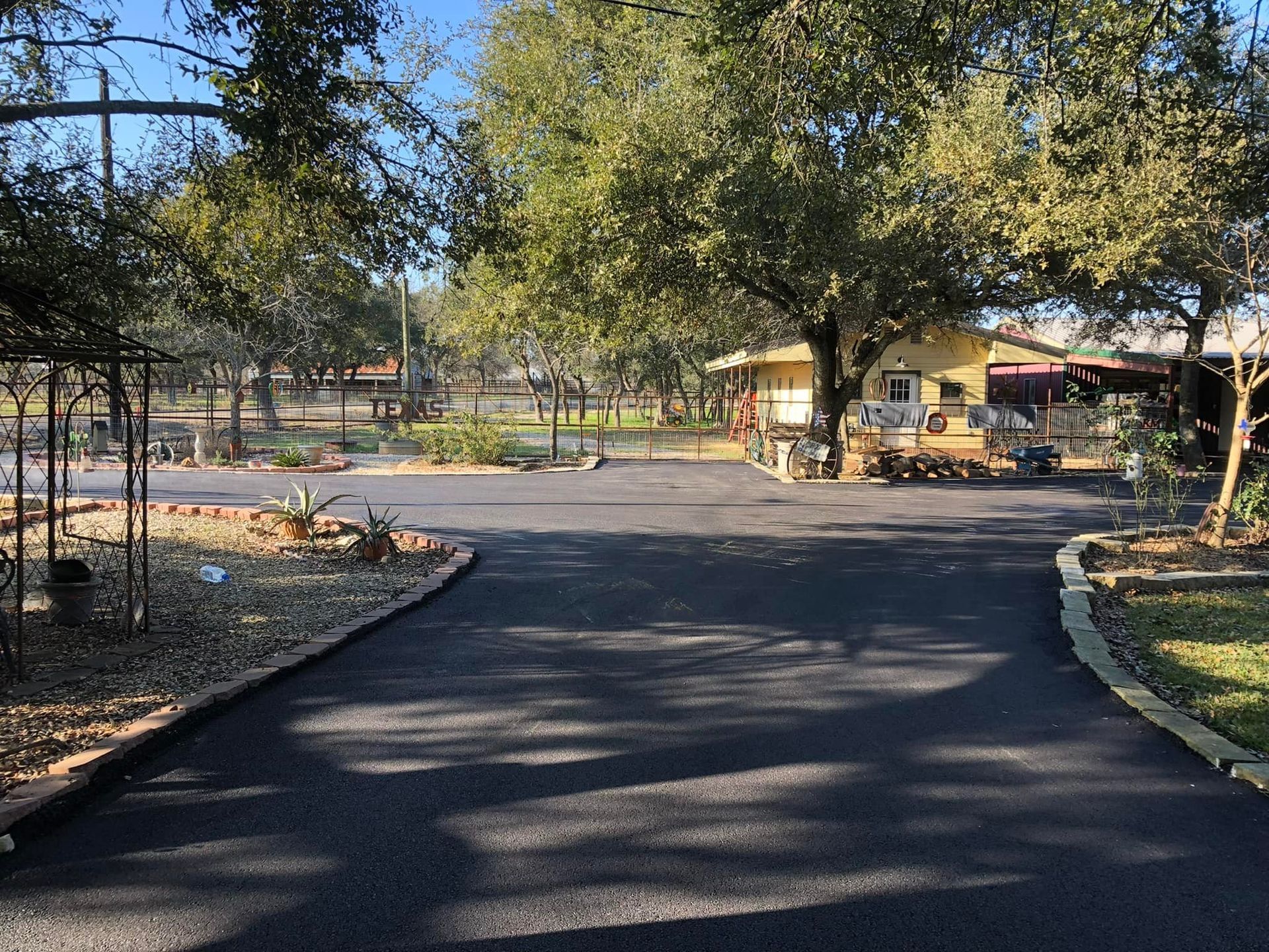 Black asphalt driveway leading to a yellow house with surrounding trees and greenery.