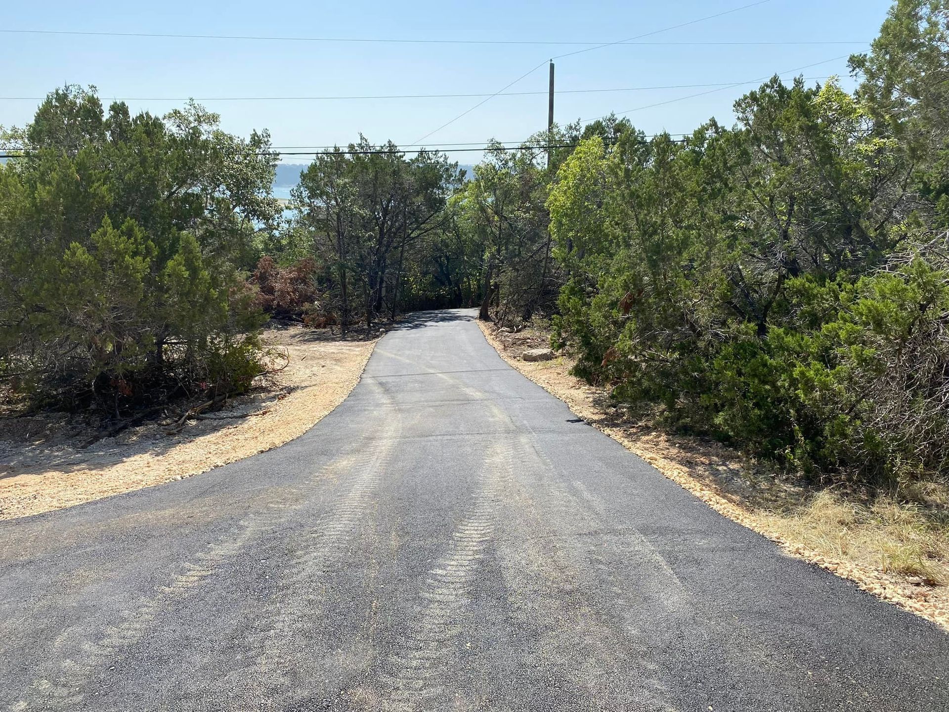 Paved driveway through trees, leading to a body of water under a blue sky.
