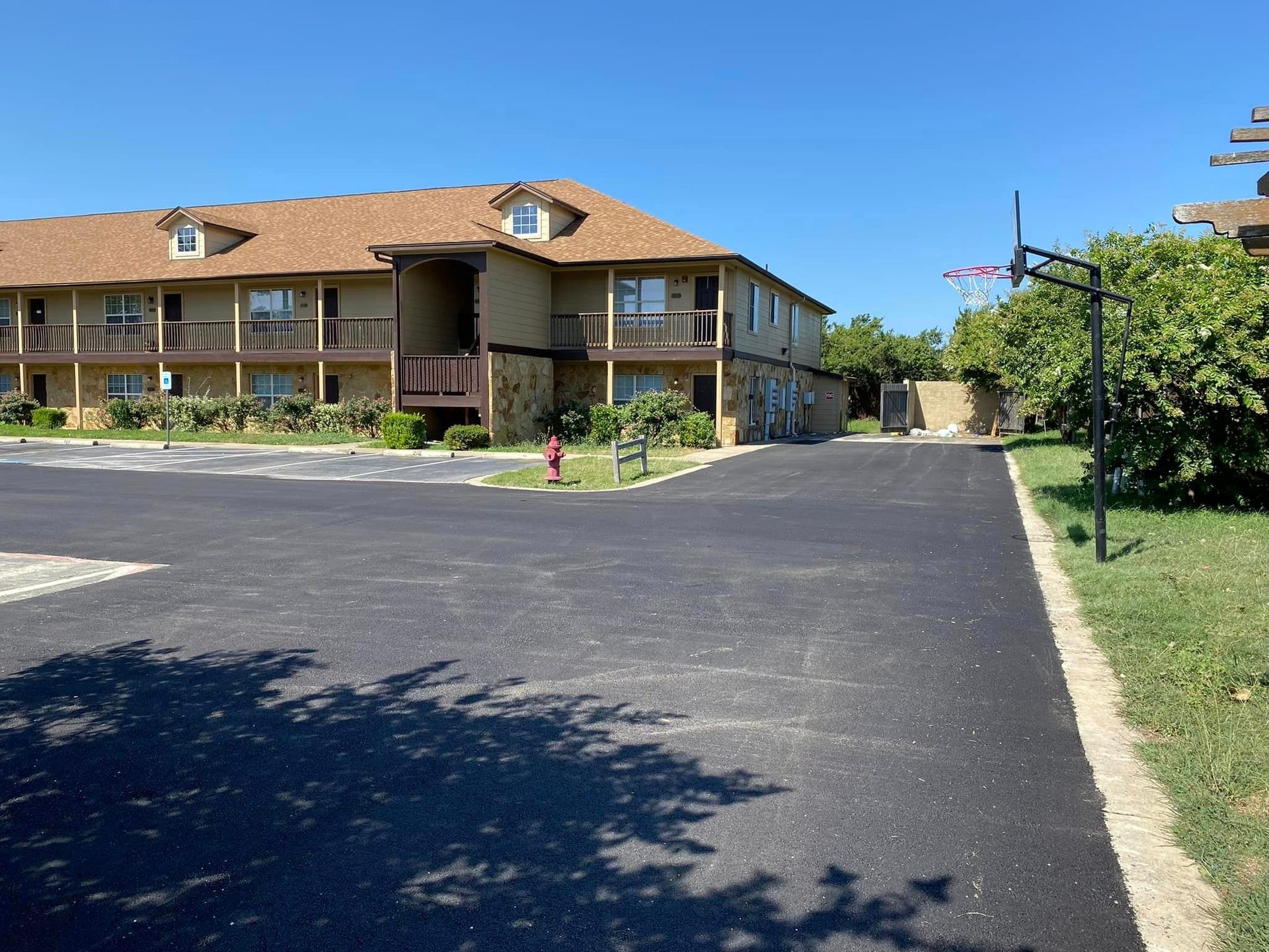 Two-story tan apartment building with balconies. Black asphalt parking lot. Clear blue sky.