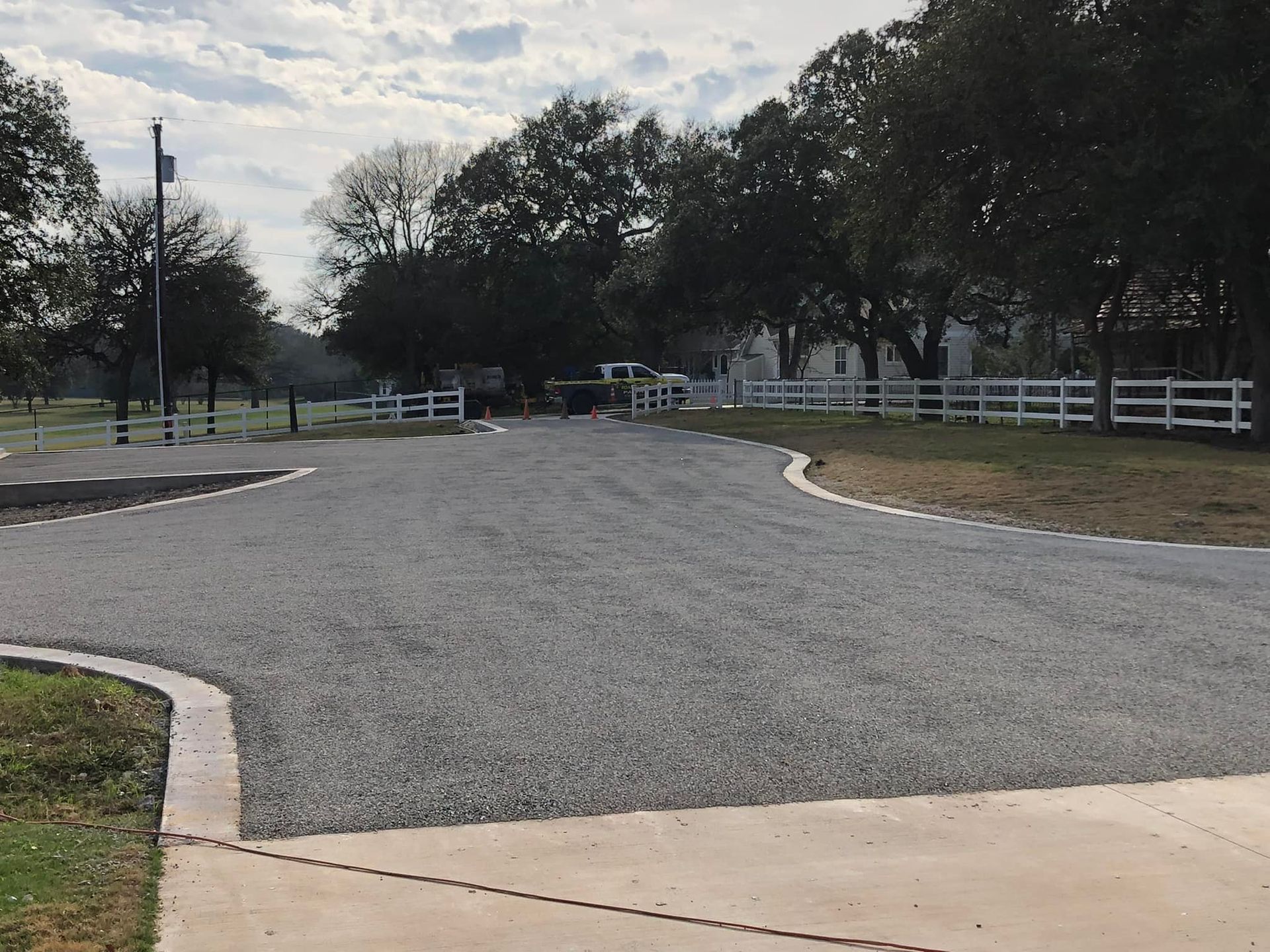 Gravel driveway curves toward white fence, trees in background. Overcast sky.