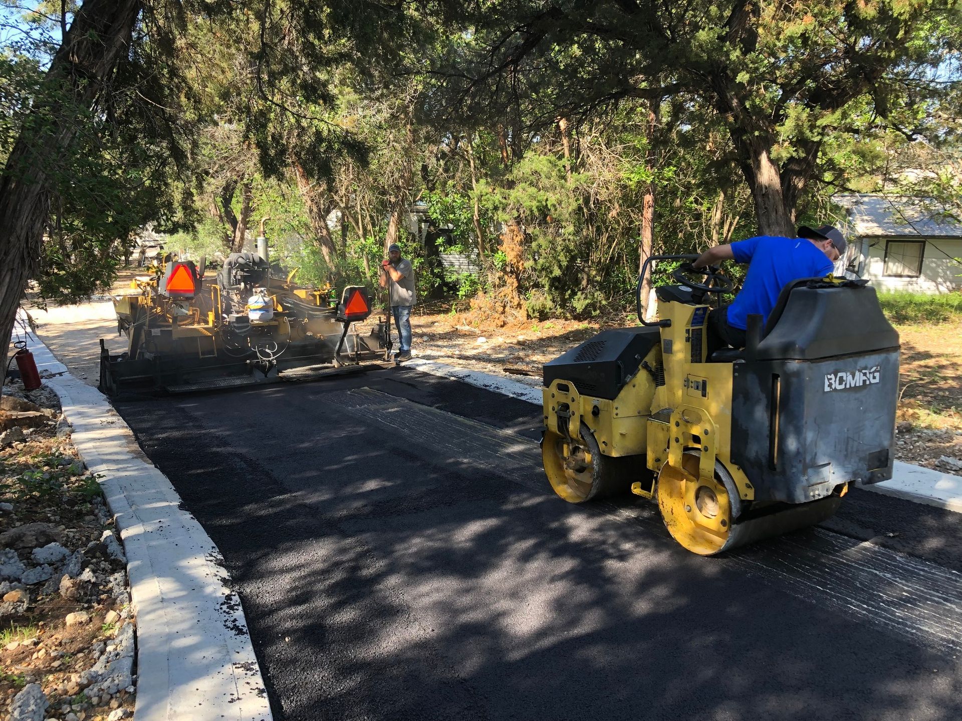 Road paving in progress: asphalt roller compacts fresh blacktop. Workers and equipment on a street.