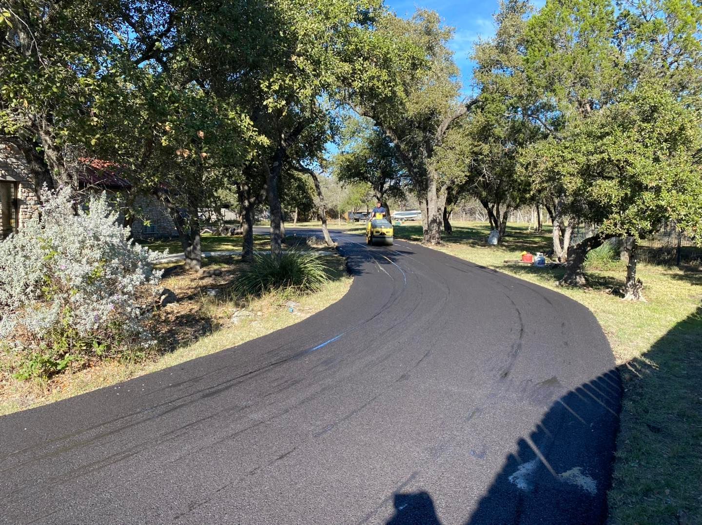Newly paved asphalt road curves through a tree-lined area on a sunny day.