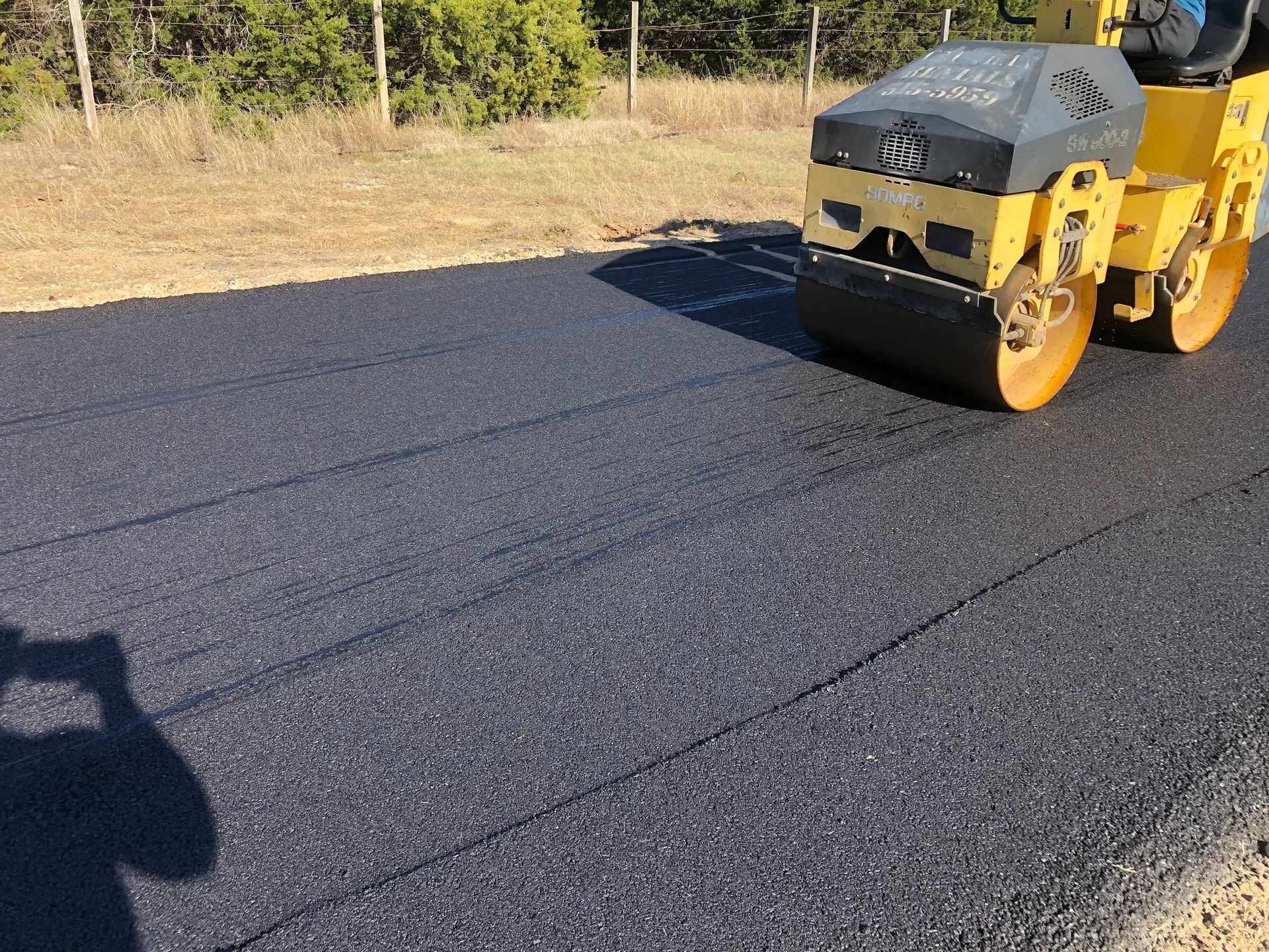 Yellow road roller compacting fresh, black asphalt on a roadside.