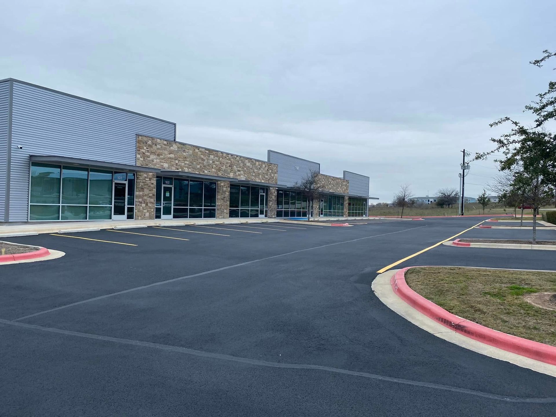 Commercial building with several storefronts, paved parking lot, and overcast sky.