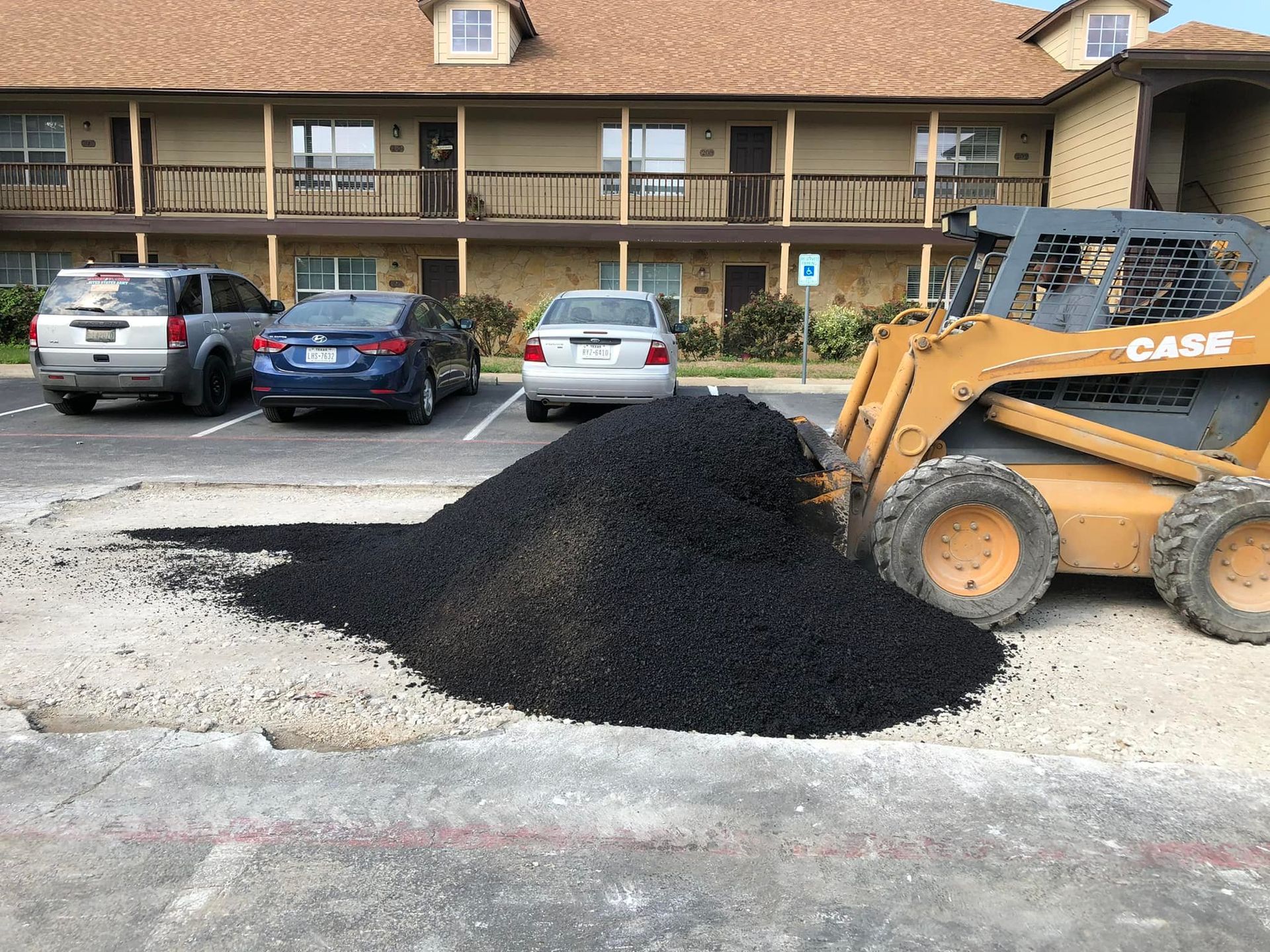 Skid steer moving a pile of black asphalt in a parking lot, near cars and a building.
