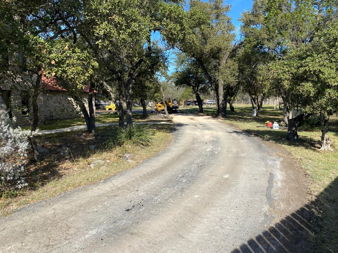 Gravel driveway curves toward a cluster of trees, with a building visible on the left, and construction equipment in the distance.