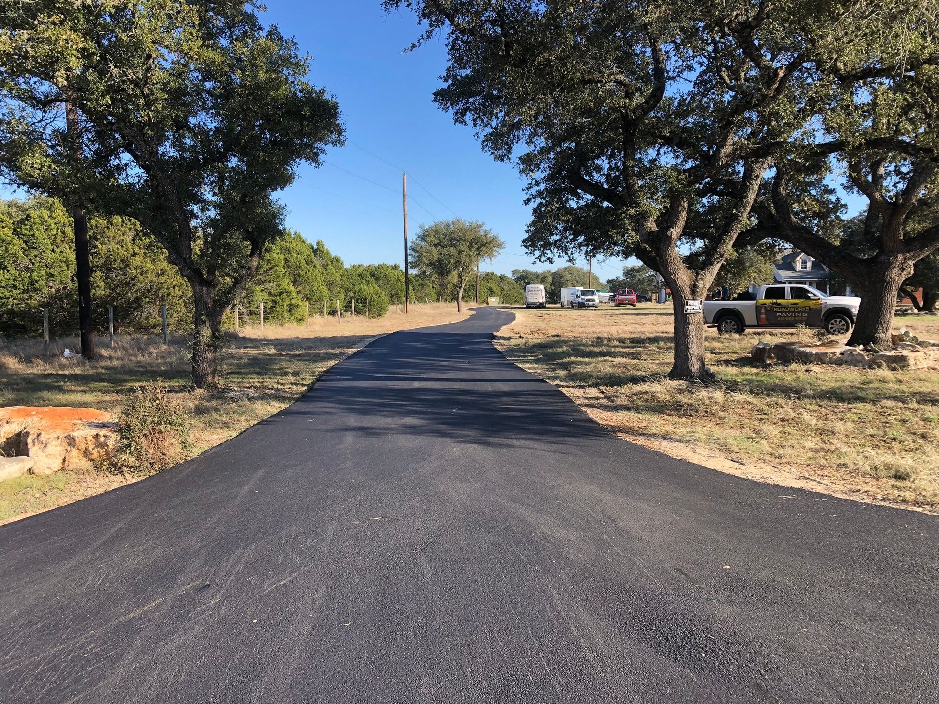 Paved road curves through a field with trees on either side, under a clear blue sky.