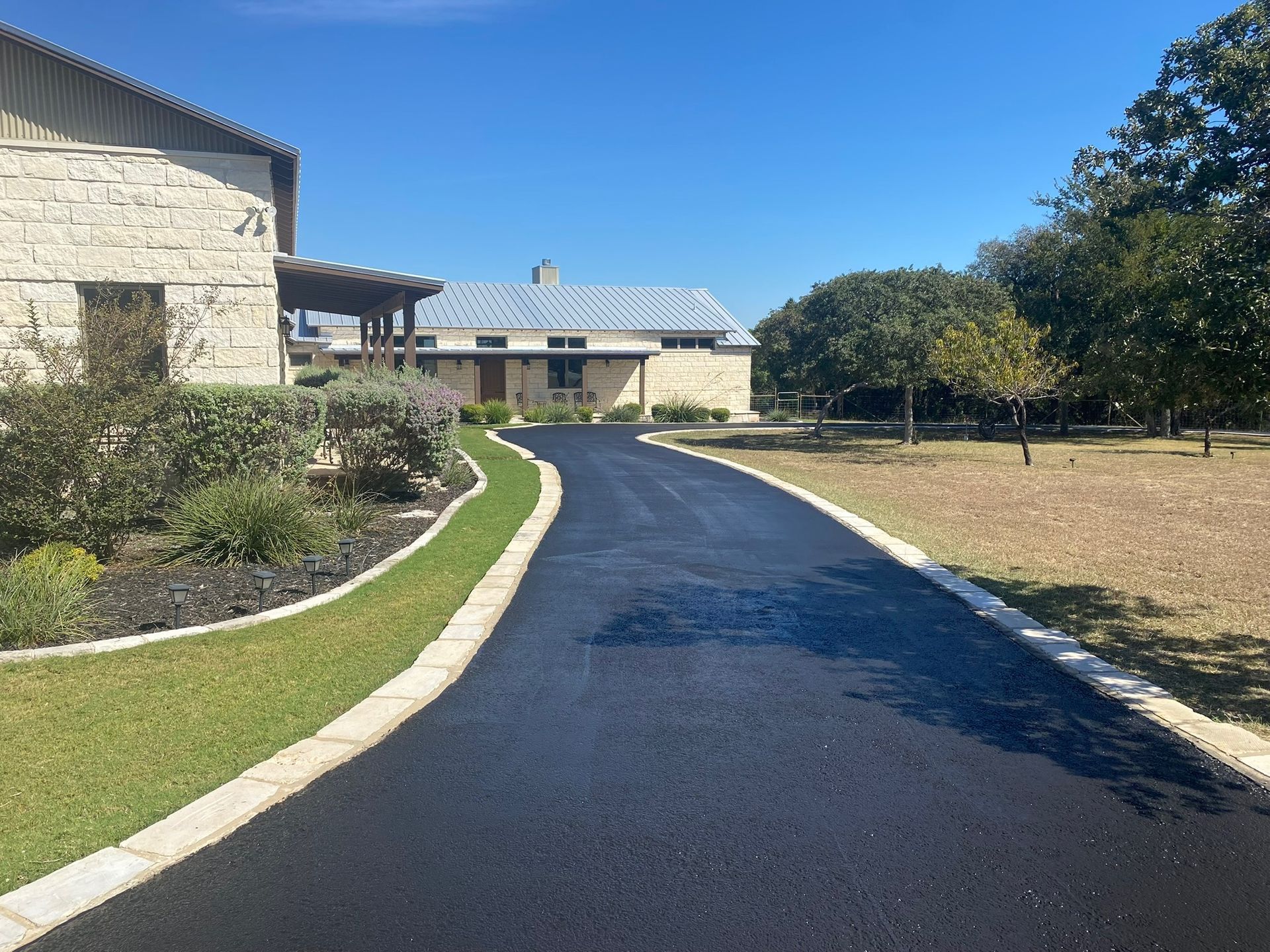 Paved driveway leads to a stone house on a sunny day, with trimmed grass and landscaping on either side.