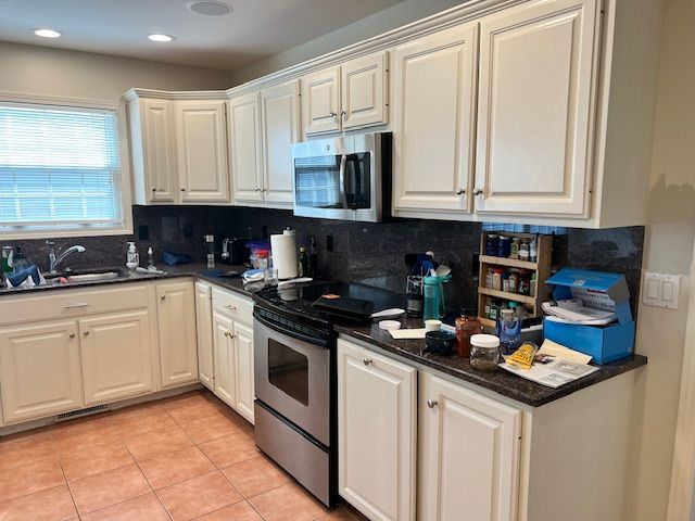 Kitchen with off-white cabinets, stainless steel appliances, dark countertops, and a window with blinds.