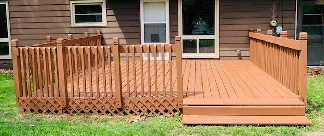 Brown wooden deck with railings, attached to a brown house with a window and a glass door, on a grassy lawn.