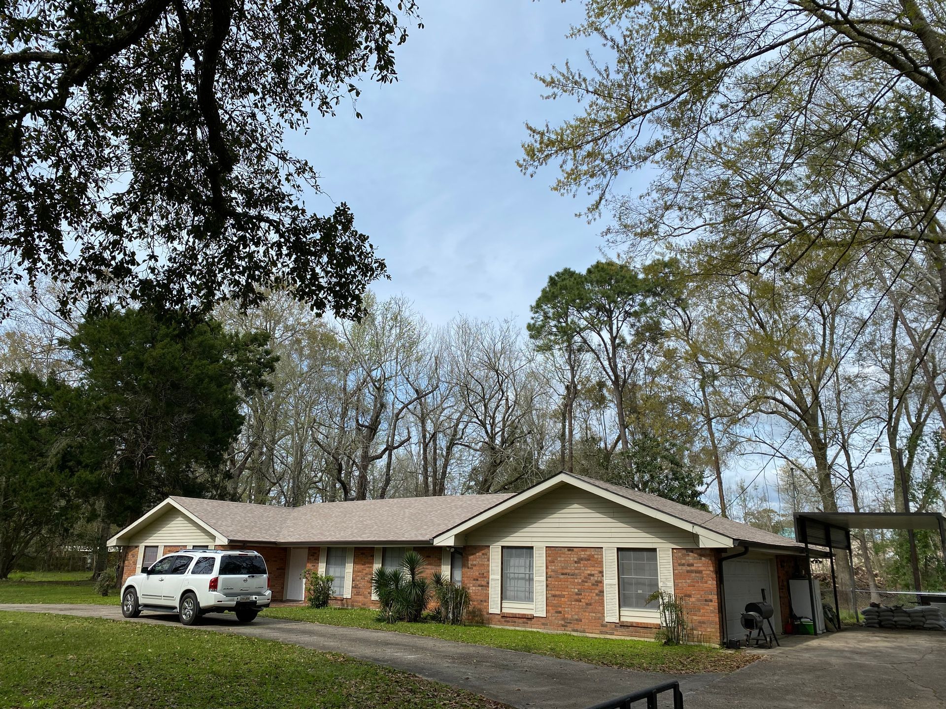 A brick ranch house with a white car parked in the driveway, surrounded by trees under a cloudy sky.
