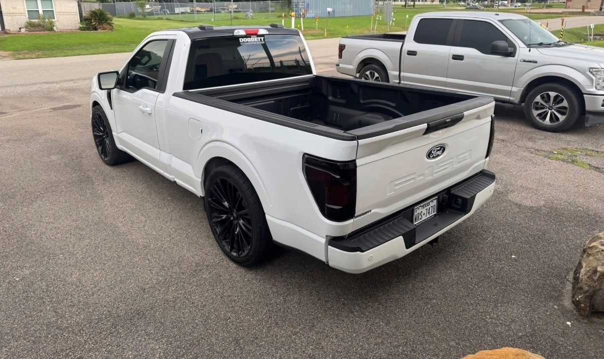 A white Ford F-150 regular cab pickup truck with black wheels and tinted taillights parked on a gravel lot.