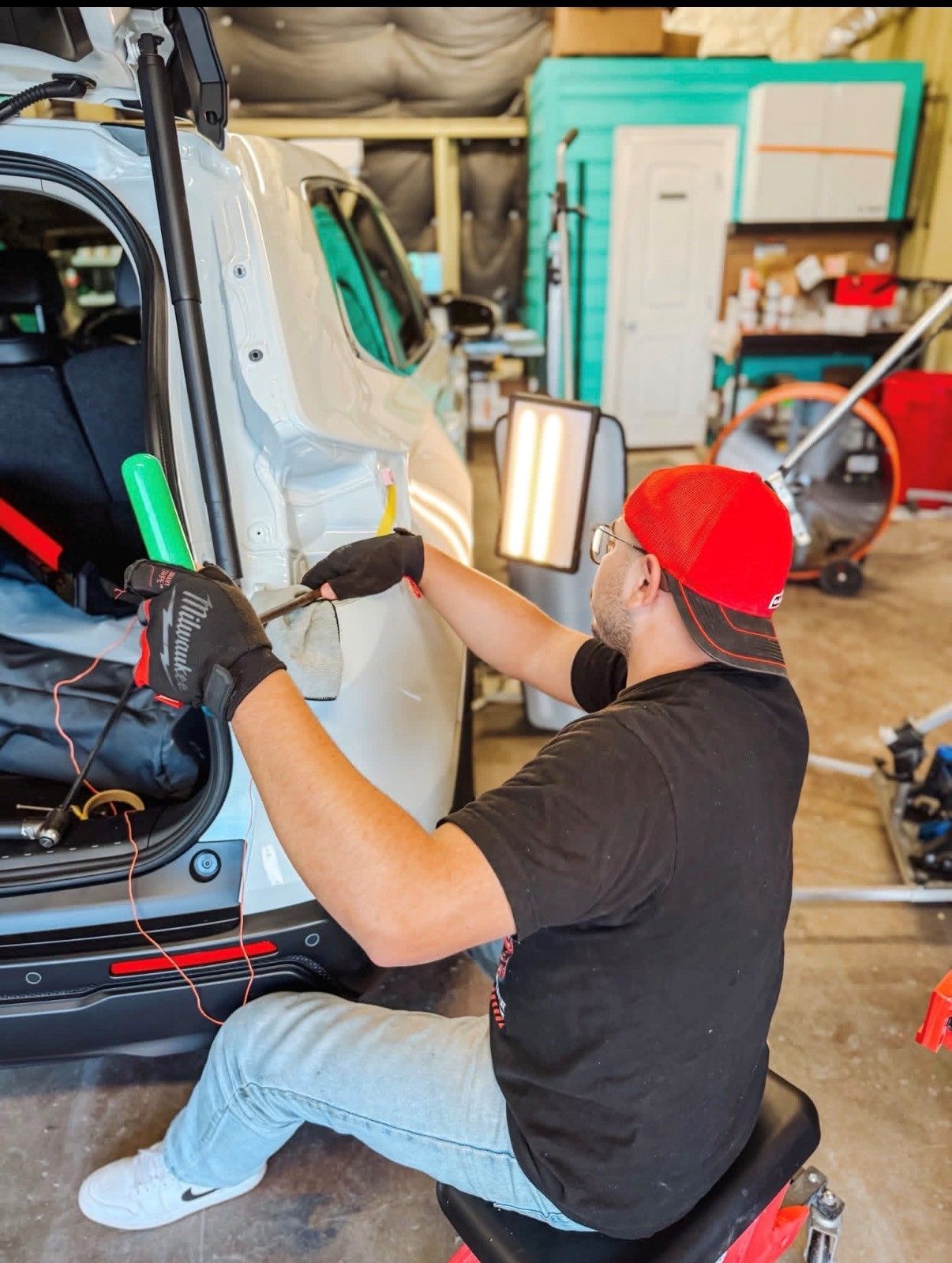 A person in a red cap and black shirt sits on a stool, performing paintless dent repair on the rear of a white vehicle.