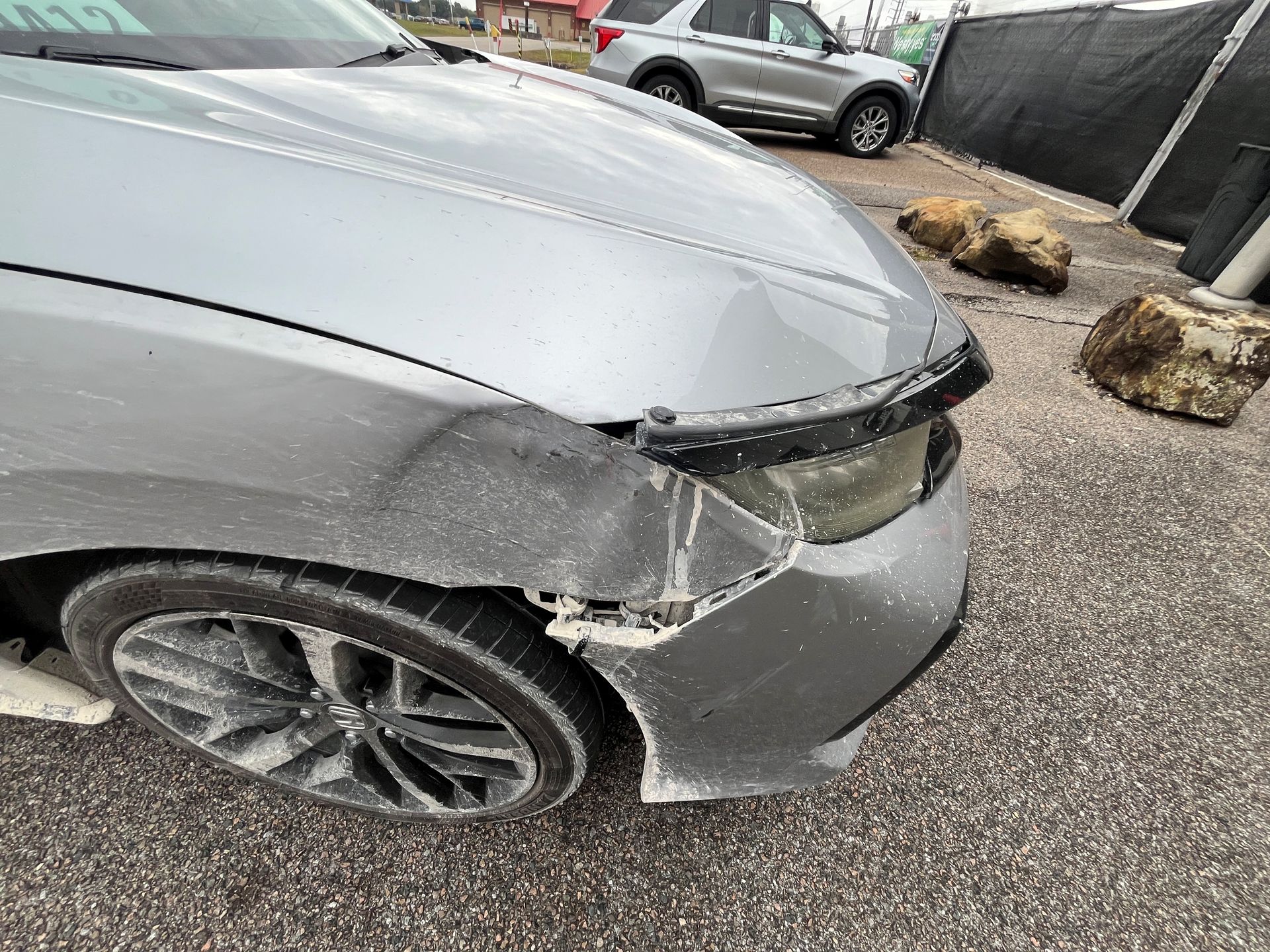 A silver car with damage to the front bumper and fender, parked on a gravel lot.