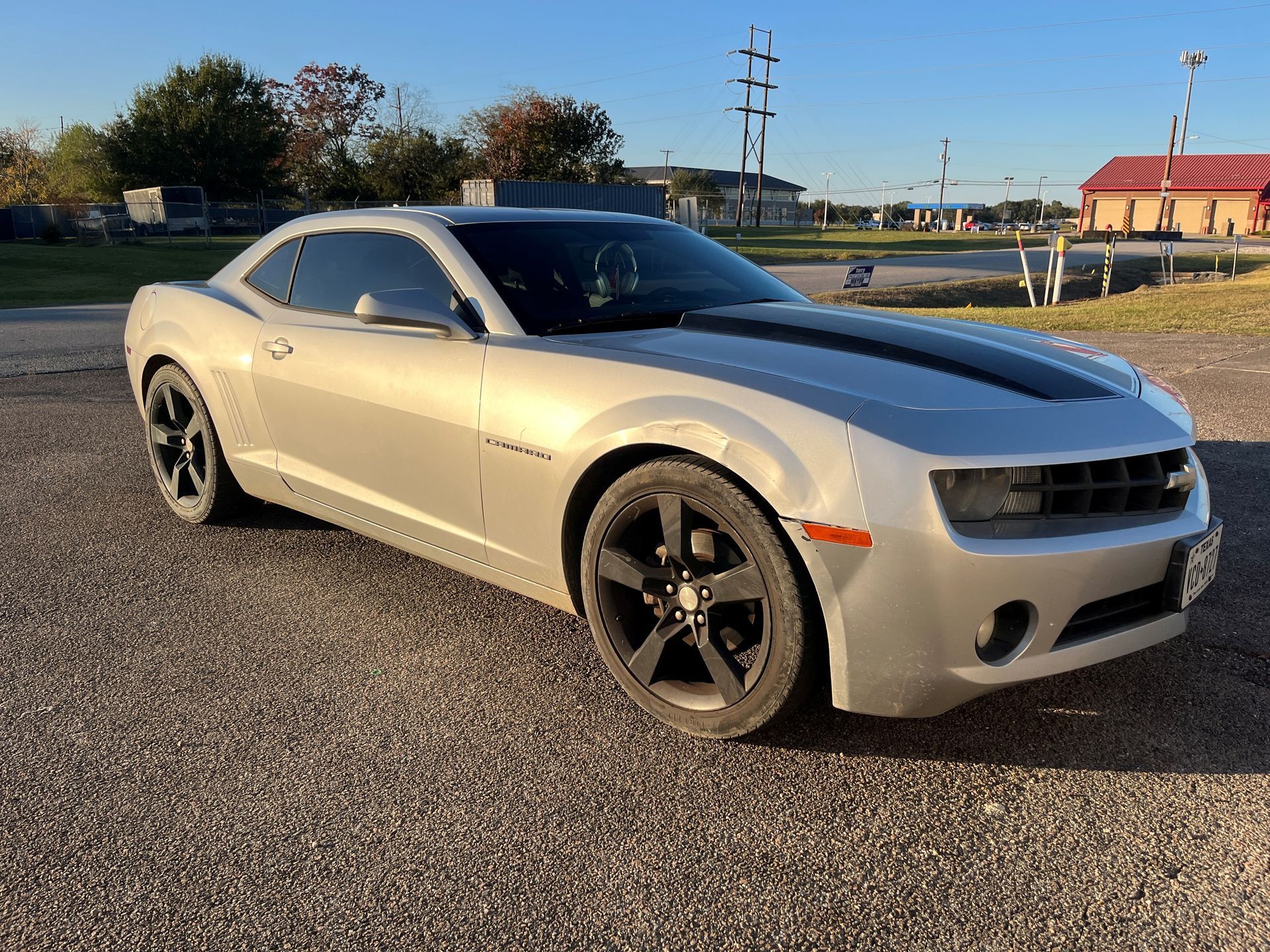 A silver Chevrolet Camaro with black racing stripes and black wheels parked on a gravel lot during the daytime.