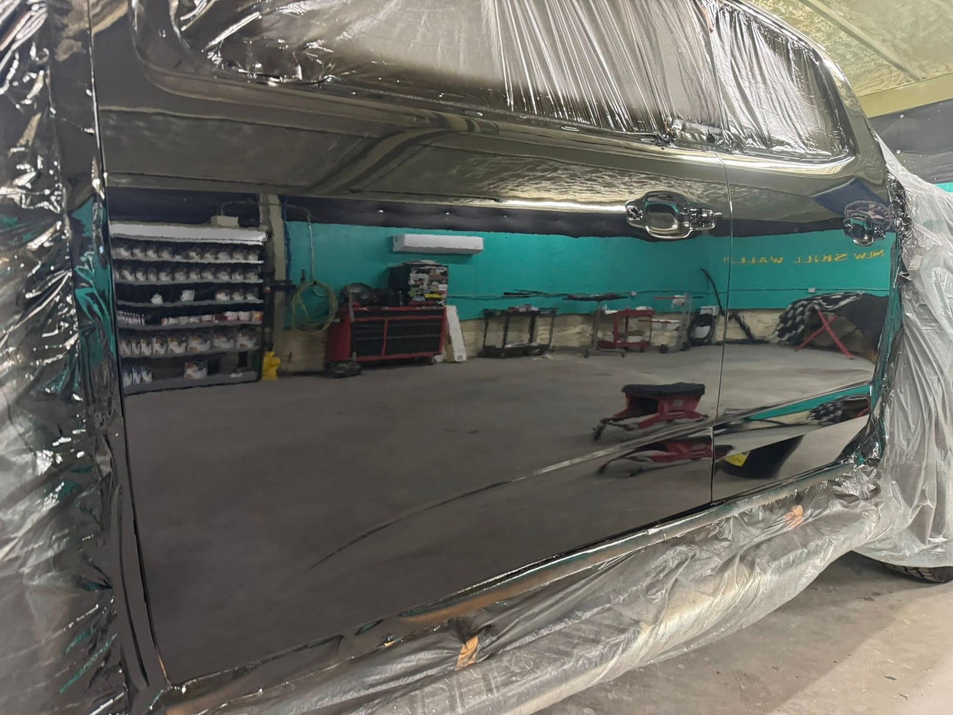 A freshly painted black vehicle door with a high-gloss finish, reflecting the interior of an auto body shop.