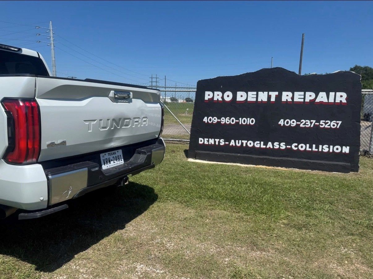 A white Toyota Tundra parked on grass next to a black Pro Dent Repair sign advertising collision and glass services.