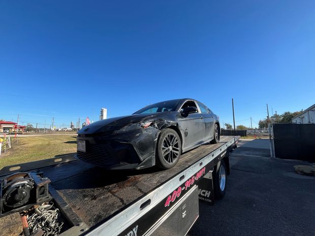 A black sedan with front-end damage secured on the bed of a flatbed tow truck against a bright blue sky.