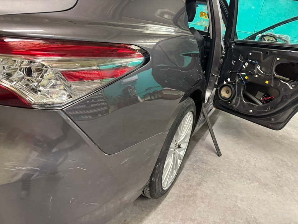 A gray car in a repair shop with its rear passenger door panel removed, exposing internal components and a speaker.