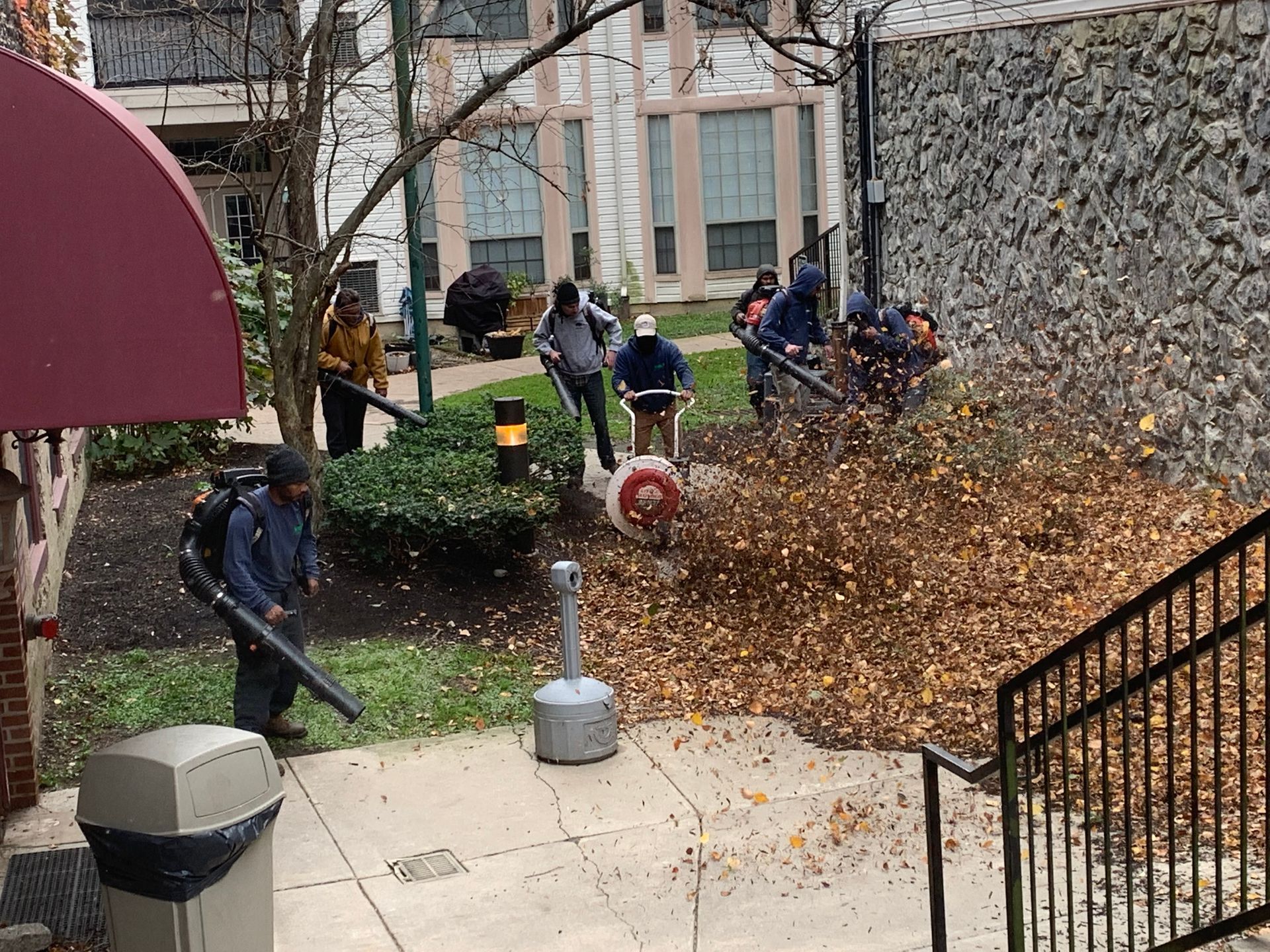 A landscaping crew uses leaf blowers to clear a large pile of autumn leaves near a building's walkway.