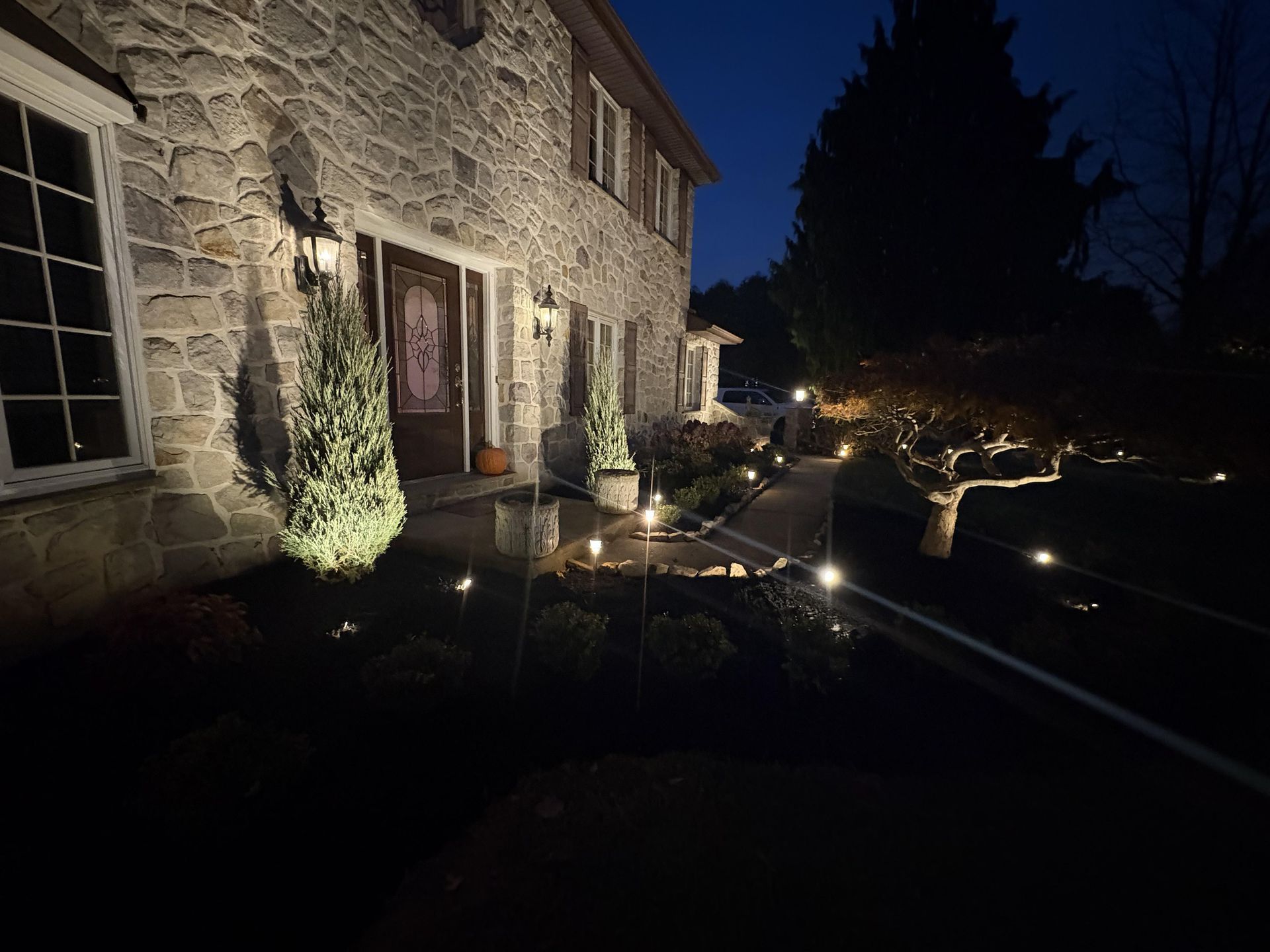 A stone house at dusk, illuminated by glowing path lights and wall sconces leading toward the front entrance.