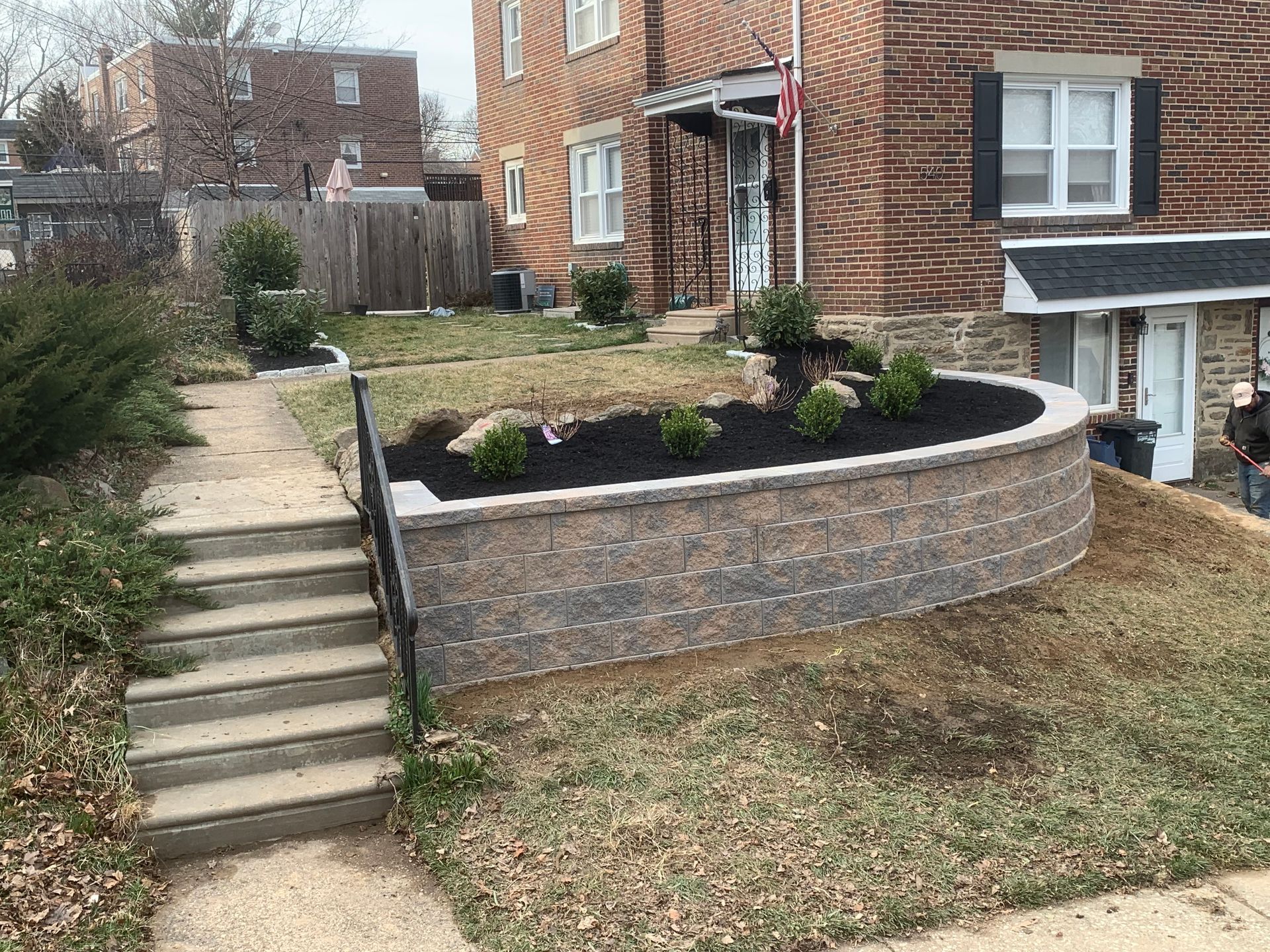 A concrete staircase leads up to a landscaped yard with a tiered stone retaining wall filled with black mulch and shrubs.