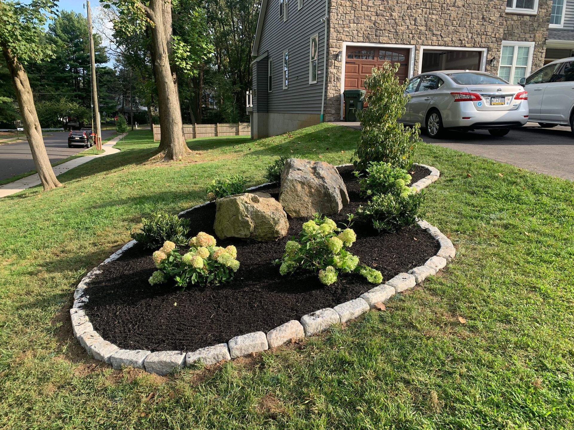 A landscaped garden bed with mulch, two large rocks, and shrubs, bordered by stone blocks on a grassy front lawn.