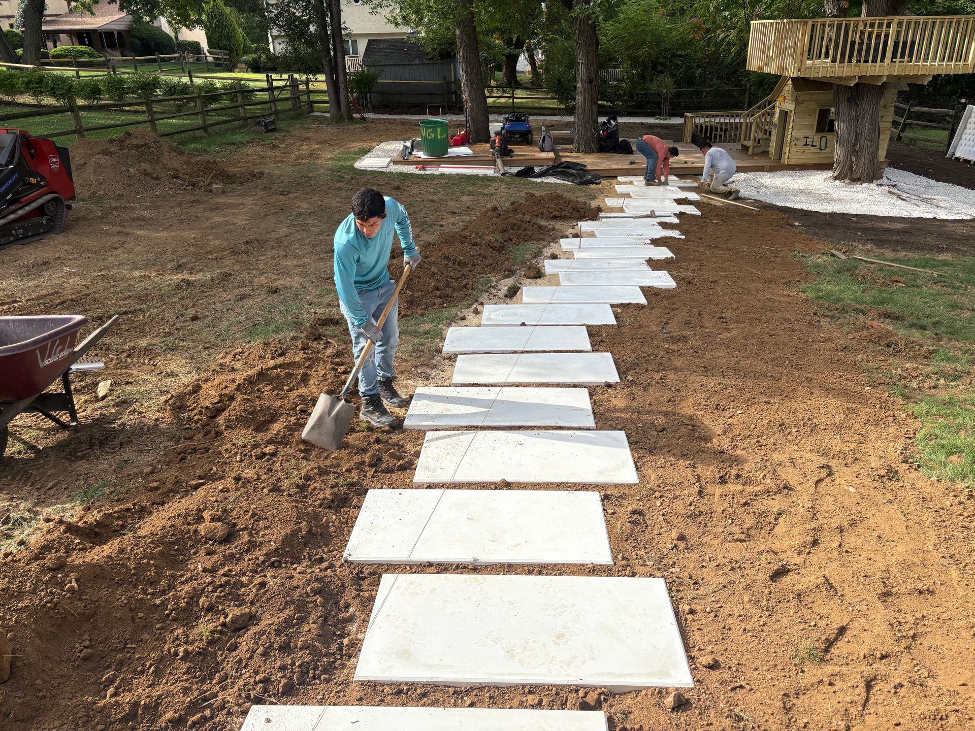 A worker shovels dirt while installing a stone path in a yard near a wooden play structure and treehouse.