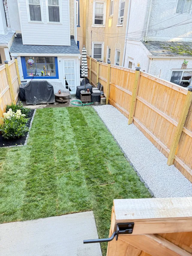A backyard view with a rectangular green lawn, a side strip of white gravel, and wooden fencing, seen from a doorway.