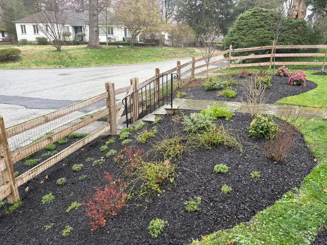 A split-rail wooden fence borders a garden bed filled with black mulch and small shrubs along a suburban street.