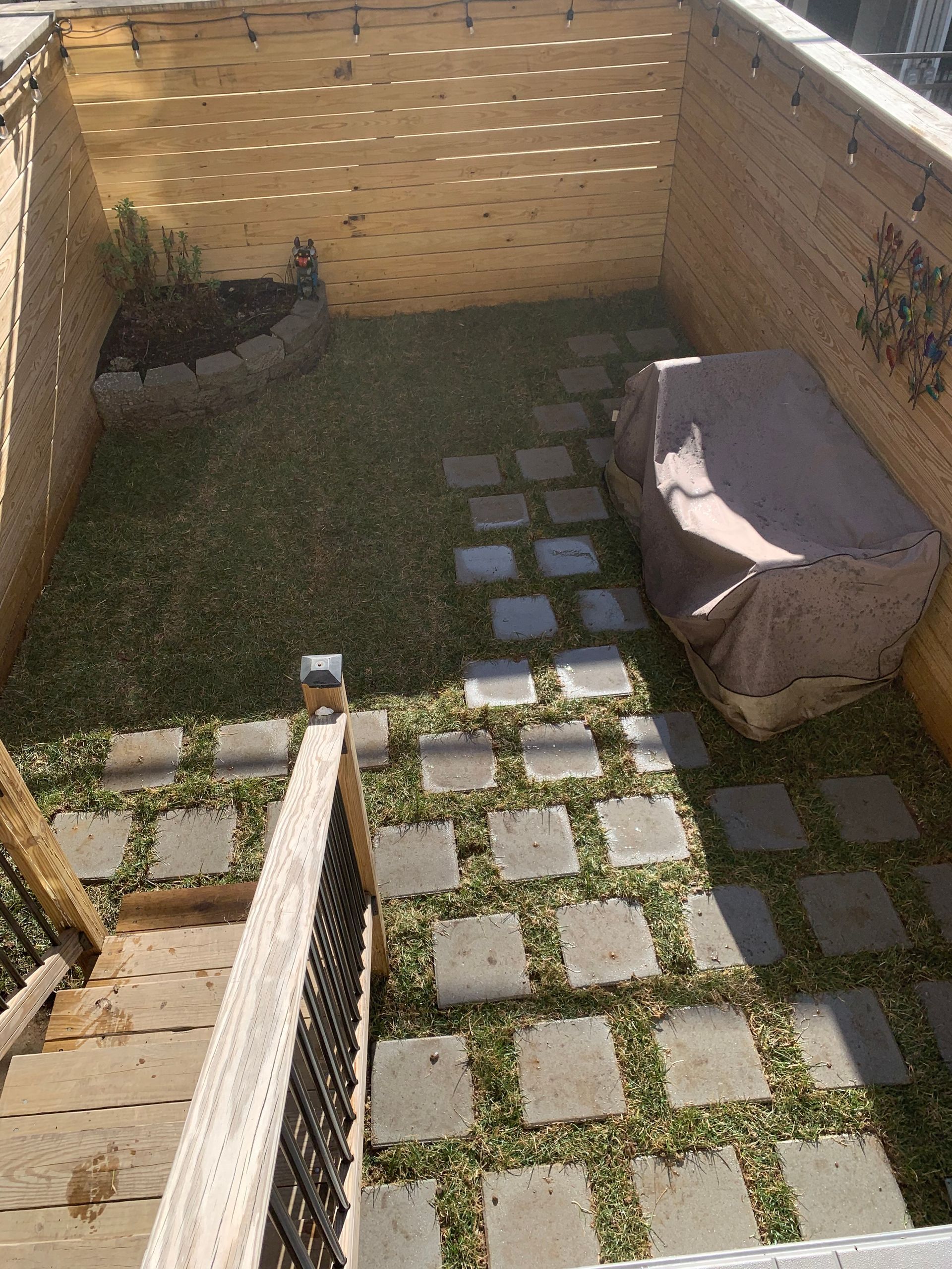 A backyard with a wooden fence, grass, a stone paver path, and a grill covered by a tan tarp, viewed from a deck staircase.