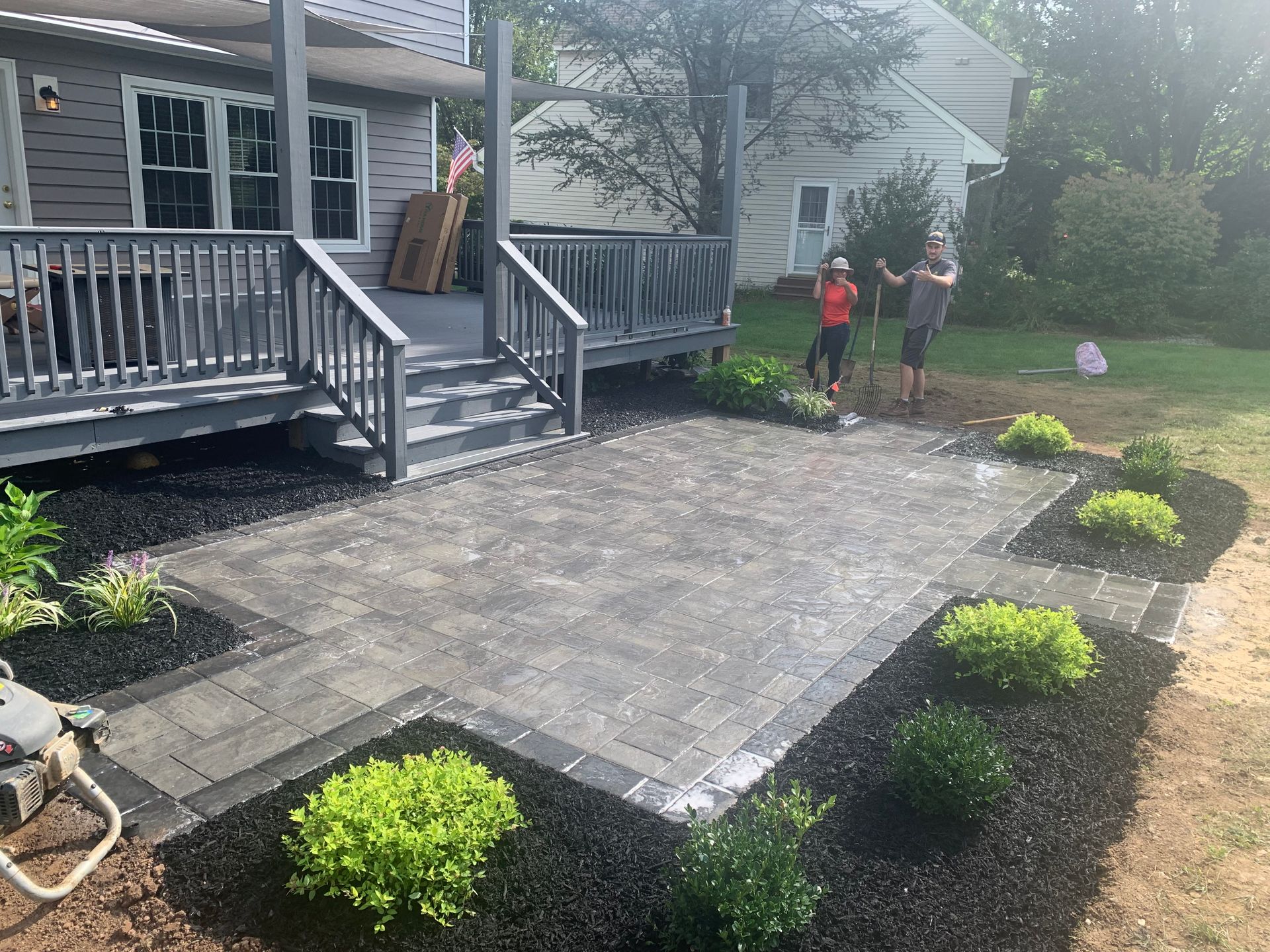 A gray paver patio sits in a backyard next to a gray wooden deck, bordered by mulch beds with small green shrubs.