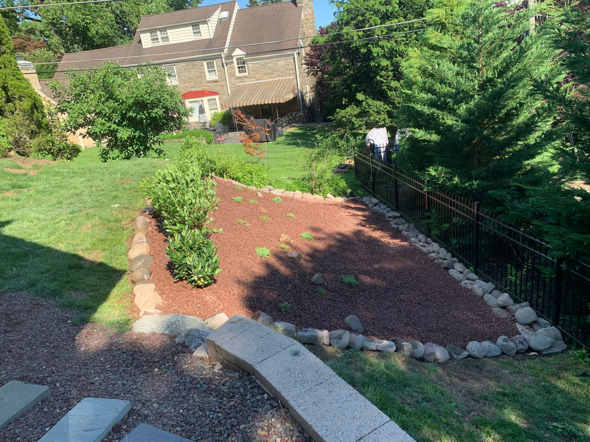 A stone-bordered garden bed with mulch and small plants in a grassy backyard next to a stone house and black fence.