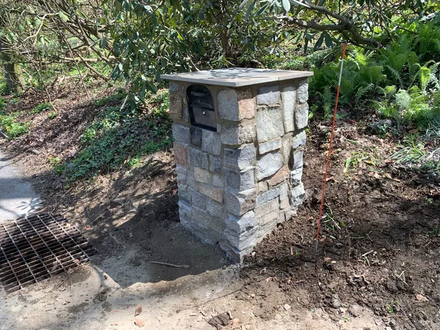 A stone mailbox pillar stands in a dirt area next to a paved path and a metal drain grate, surrounded by trees and brush.