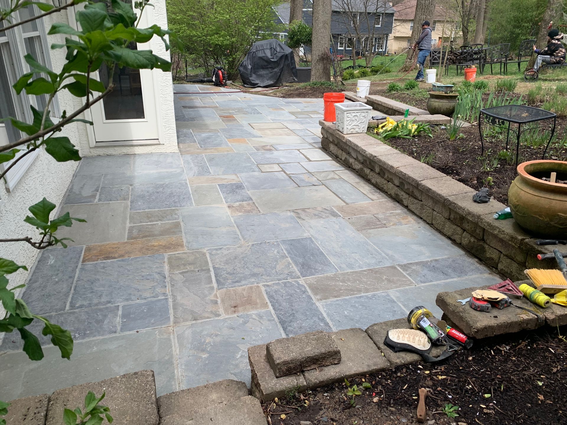 A newly installed patio of rectangular gray and tan stone pavers next to a house with a low retaining wall.