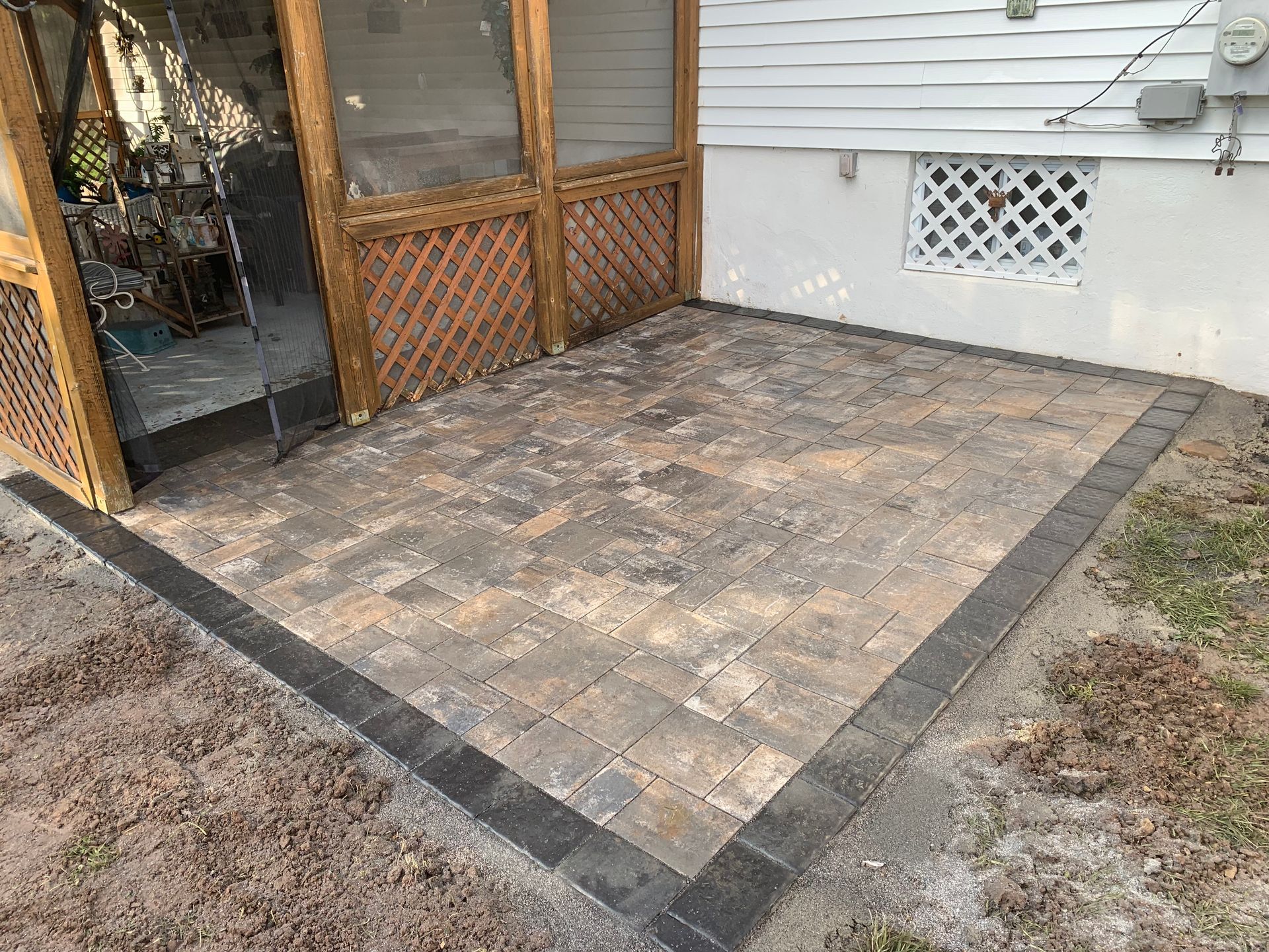 A new paver patio with a dark border sits next to a home with white siding and a screened porch.