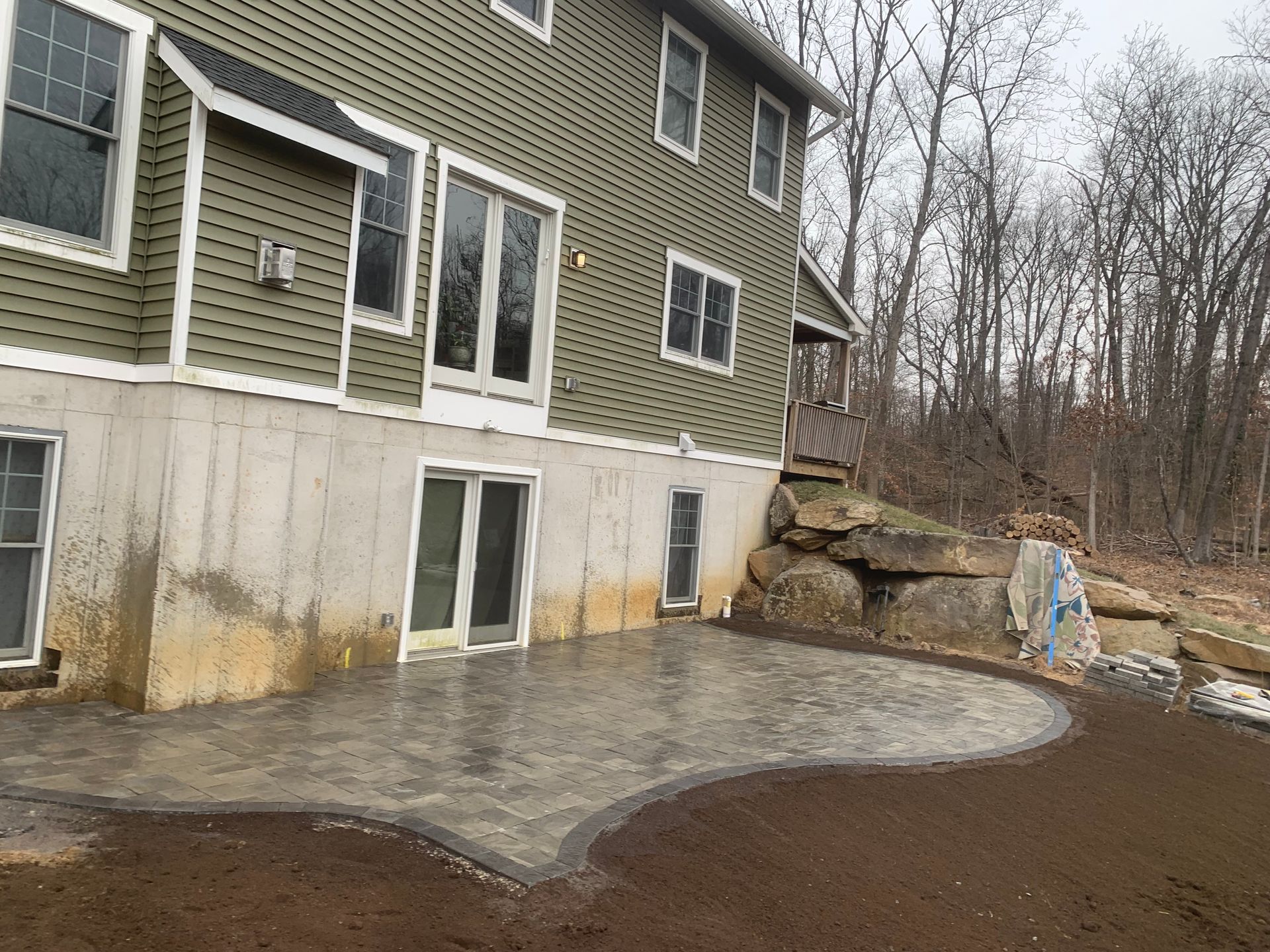 A newly installed stone paver patio sits against the concrete foundation of a green-sided house, next to a rock wall.