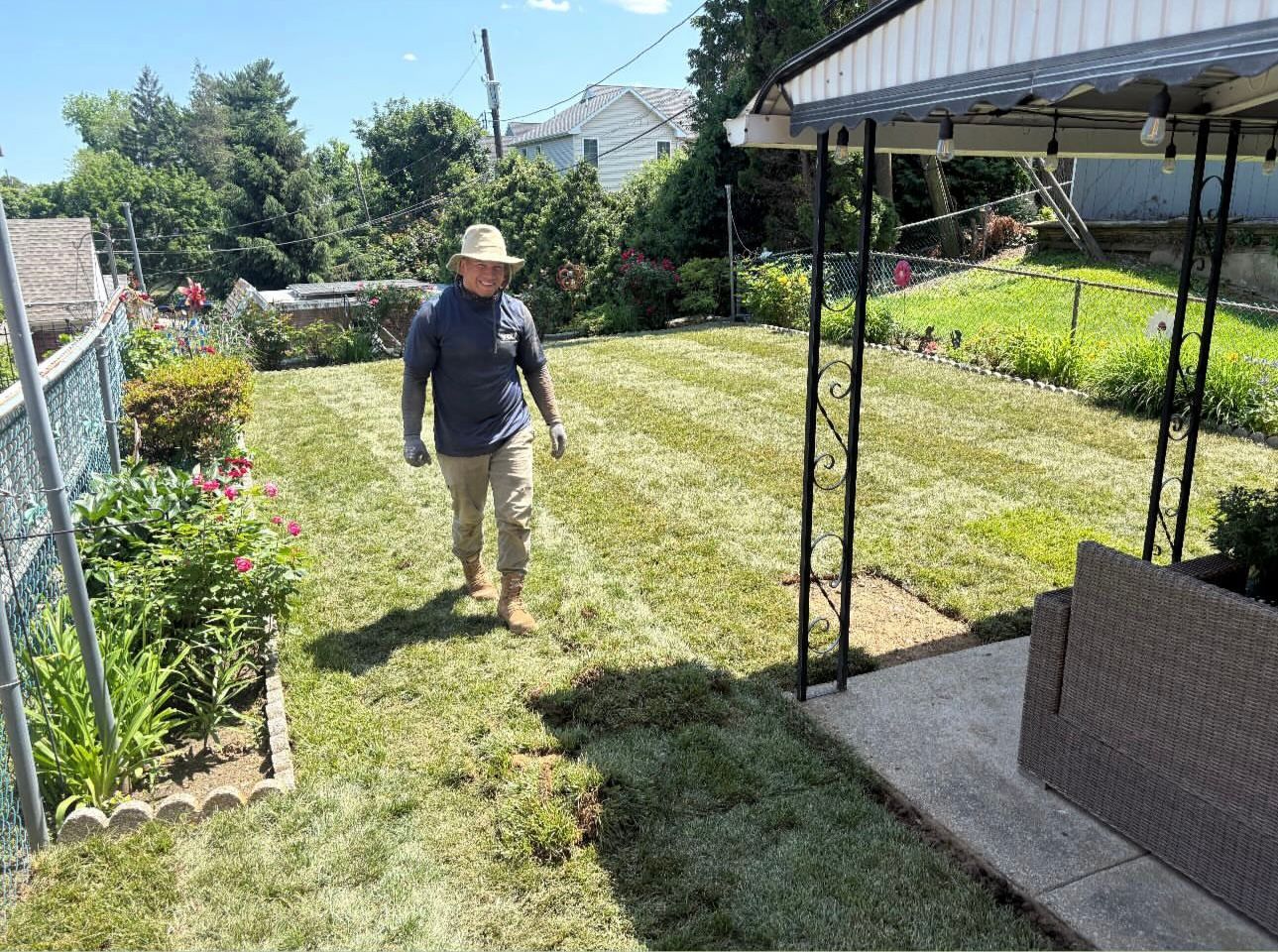 A person wearing a straw hat and dark shirt walks across a recently mowed backyard lawn near a patio.