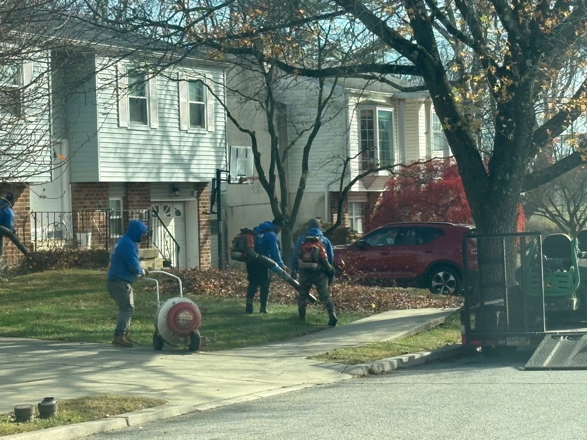 Workers in blue hoodies use leaf blowers and a wheeled vacuum to clear autumn leaves from a residential front yard.